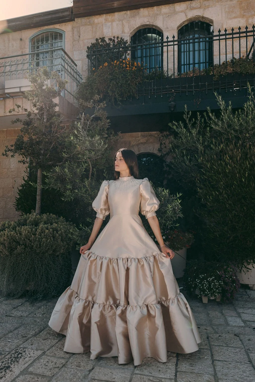 A woman in an elegant cream-colored gown with puffed sleeves stands outdoors among lush green plants, with a stone building featuring balconies and arched windows in the background.