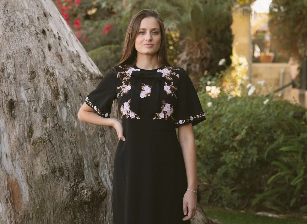 A young woman standing outside next to a large tree, wearing a black dress with floral embroidery on the top and sleeves, with a bow detail on the neckline, during daytime.