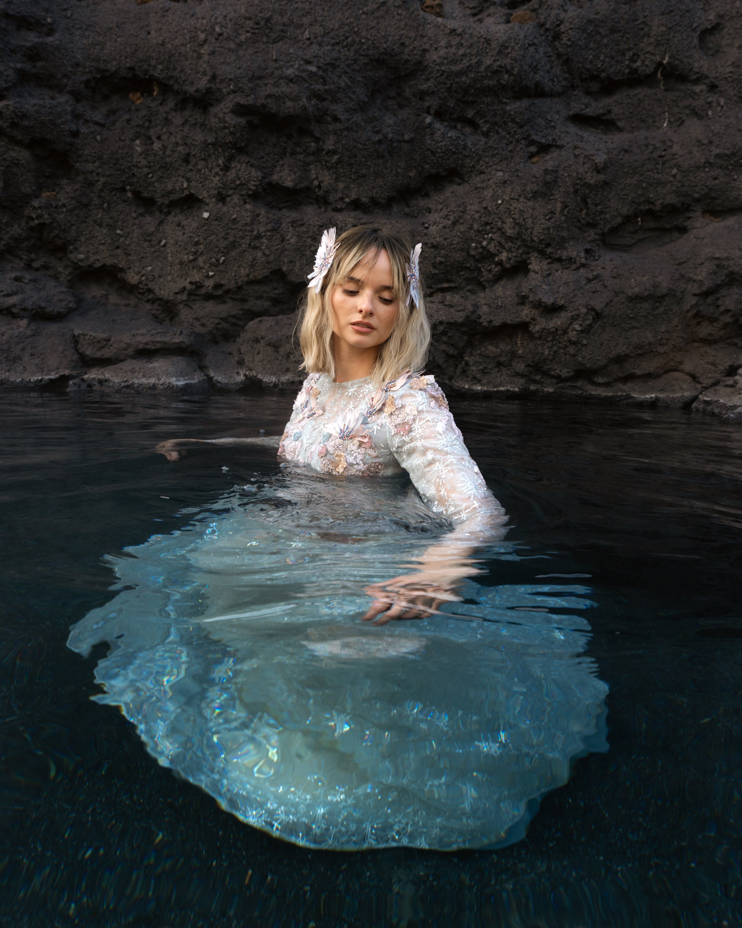 A woman with blonde hair, wearing a lace dress and feather hair accessories, stands in dark water near a rocky black shoreline, with her eyes closed and one arm extended underwater.