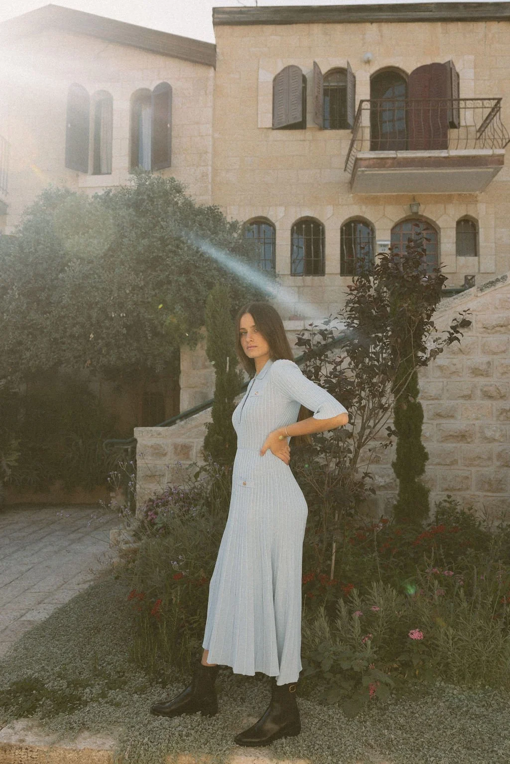 A woman in a light-colored, pleated, long dress and black boots standing outdoors near a garden and a stone building with arched windows and wooden shutters.