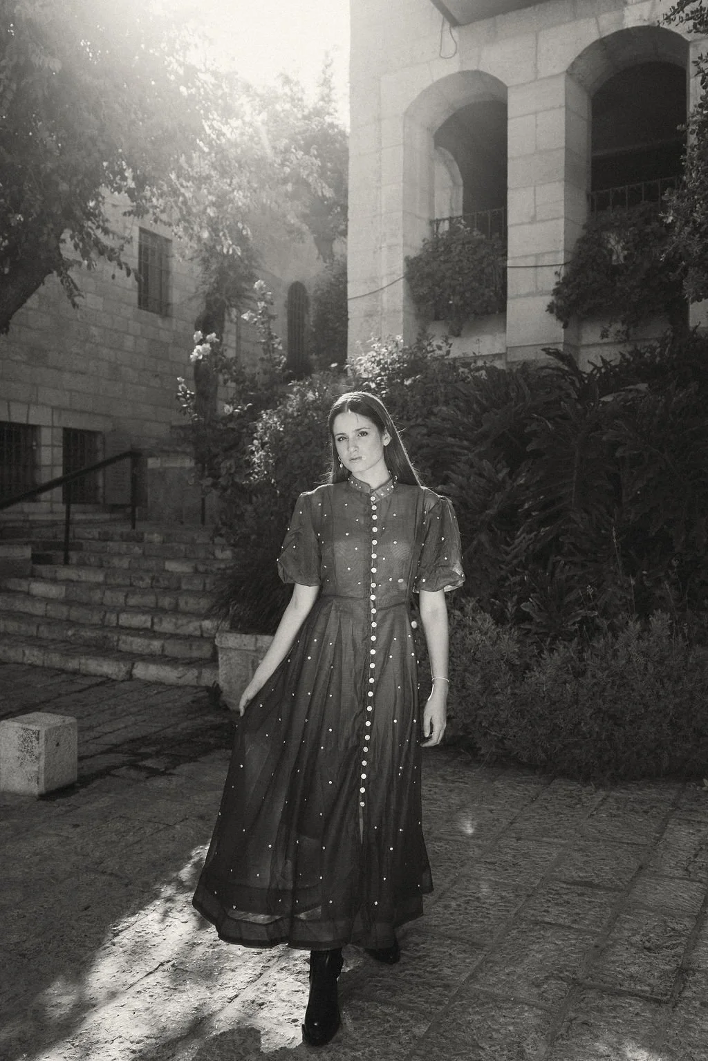 A woman standing outdoors on stone steps, wearing a long dark dress with pearl buttons, in a setting with trees and an old stone building with arched windows, during sunlight.