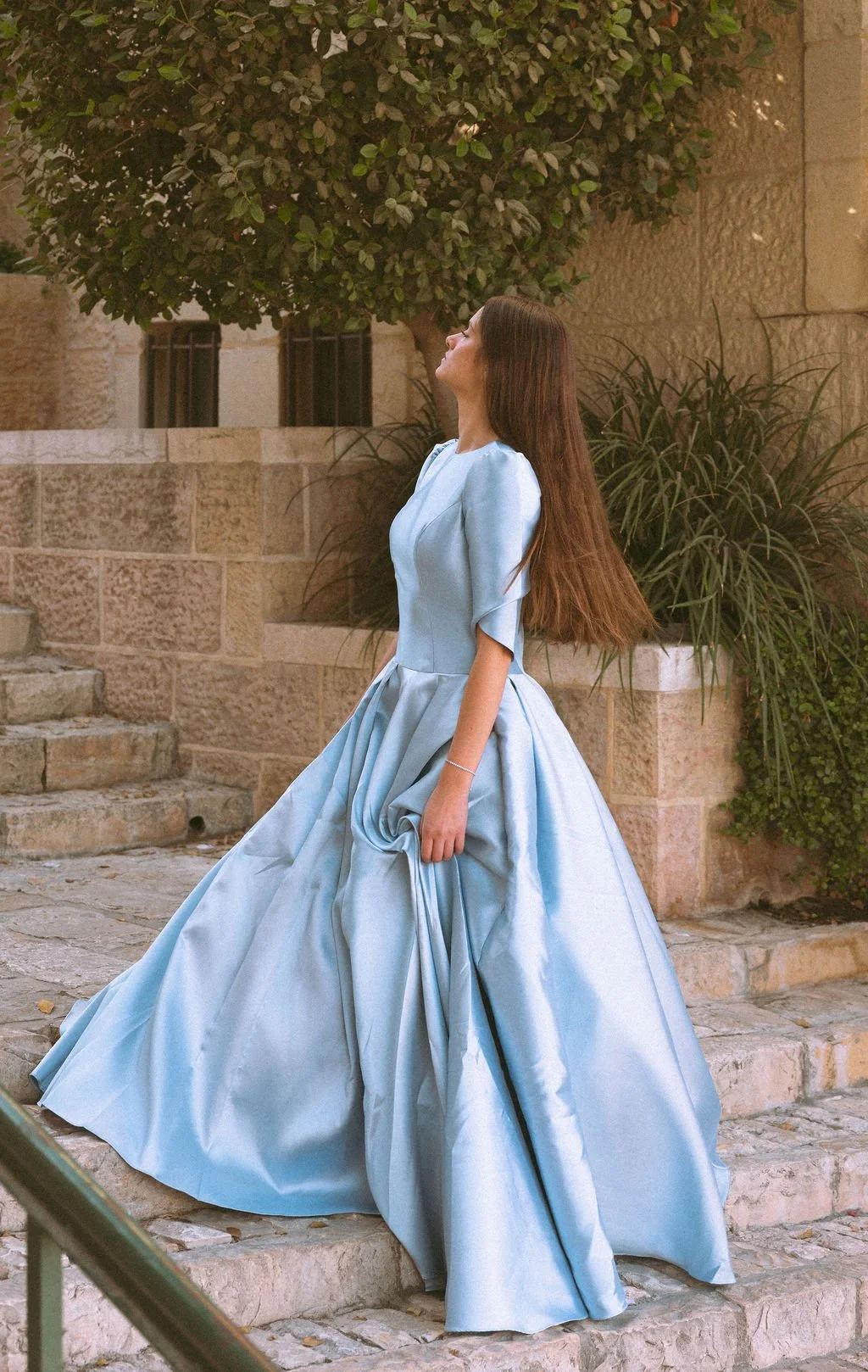 A woman in a flowing light blue gown standing on stone steps outdoors, with her head tilted back and hair flowing, near a stone wall and greenery.