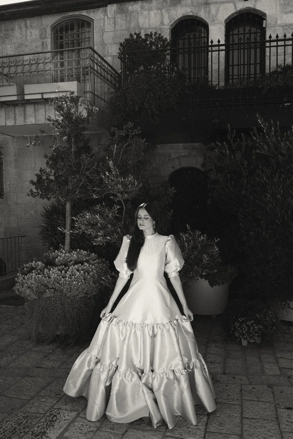 A woman in a vintage-style dress standing outdoors at night, surrounded by plants and stone architecture.