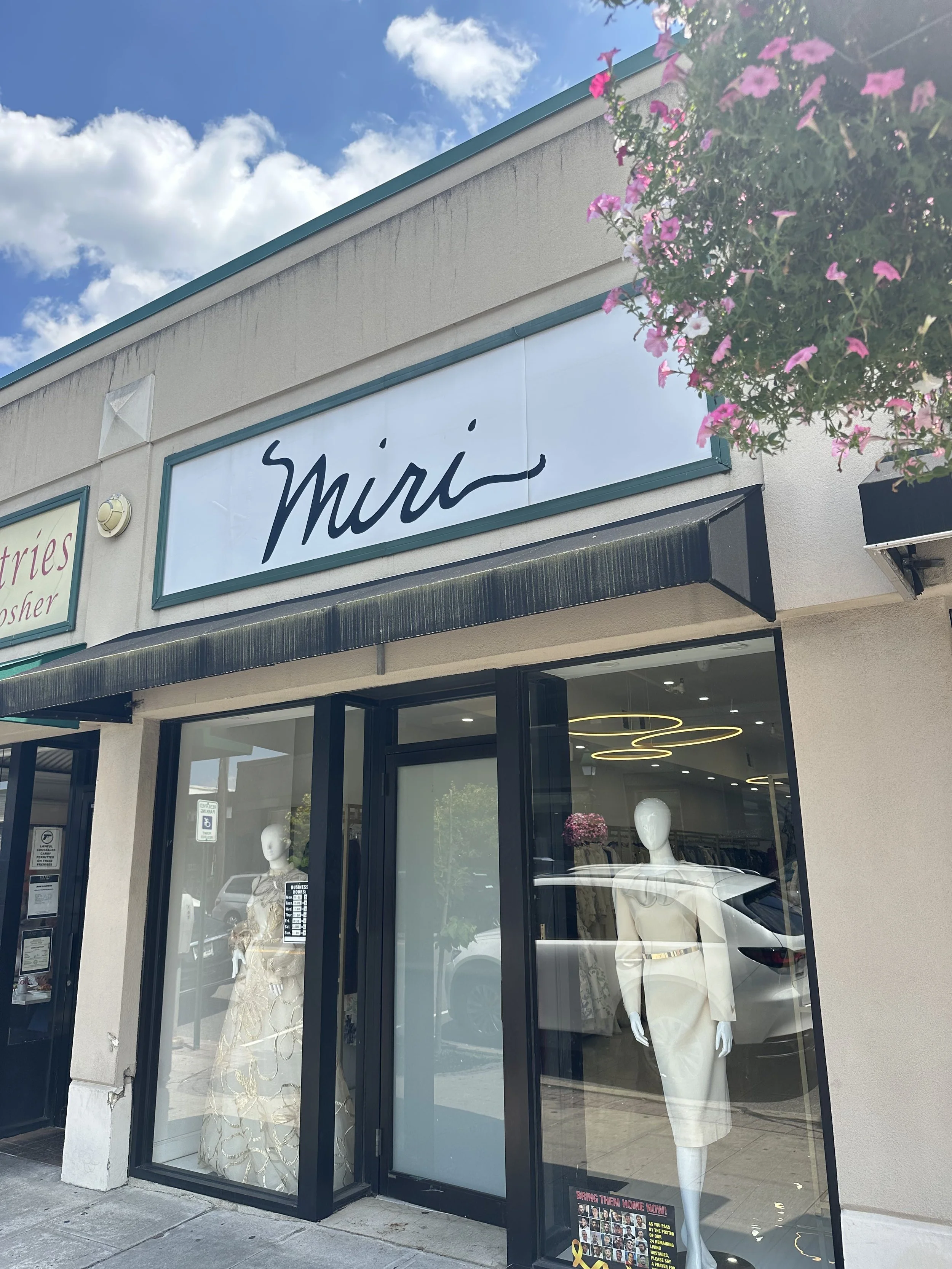 Fashion boutique storefront with mannequins dressed in elegant clothing displayed inside, glass windows, and a sign that reads 'Miri' above the entrance. A flowering plant hangs outside on the right side of the storefront.
