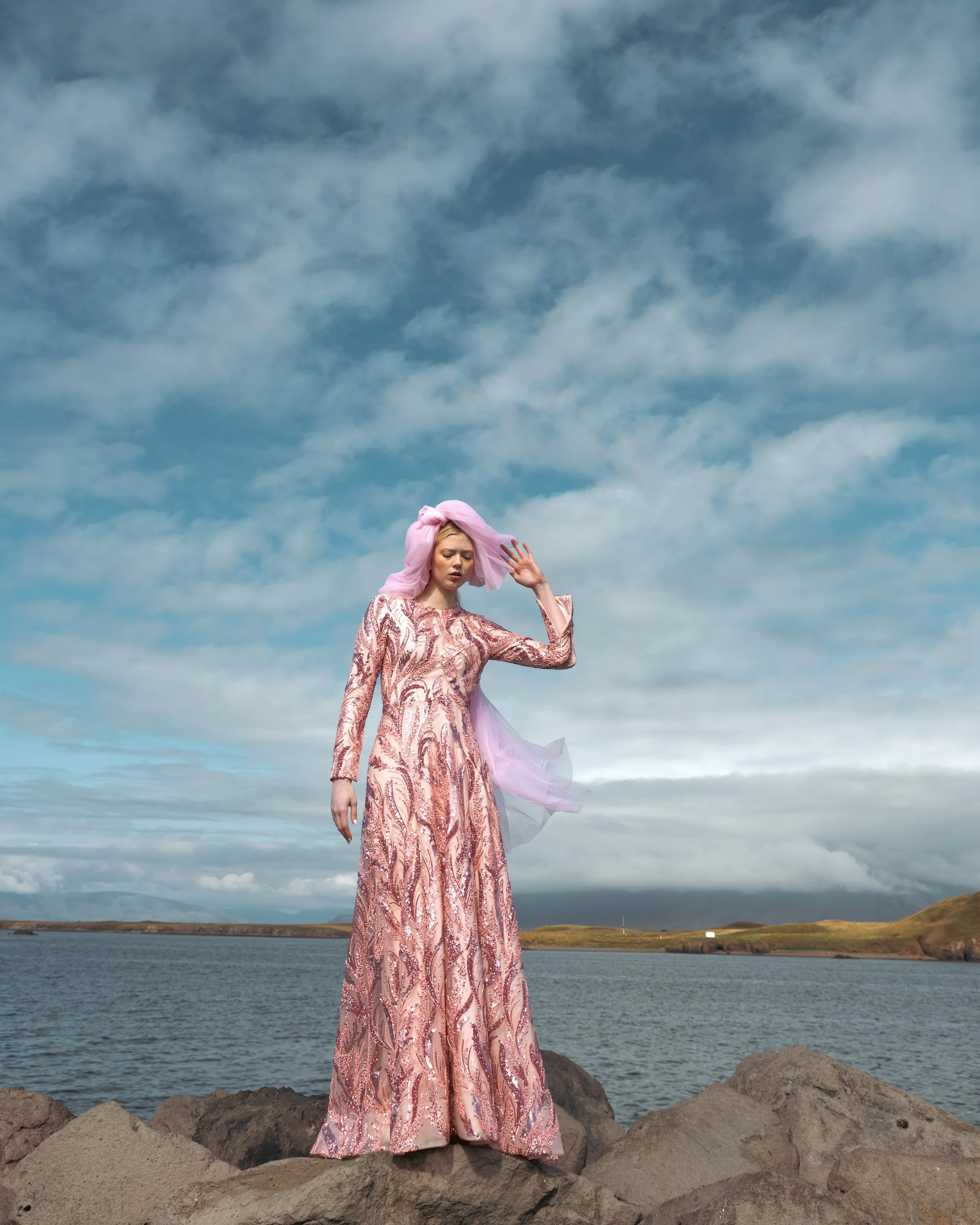 A woman in a shiny pink and rose patterned gown standing on rocks near a body of water, with a cloudy sky in the background.