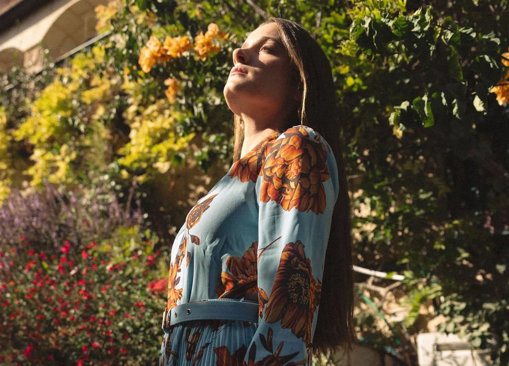 Woman with long hair in a floral dress standing outdoors in sunlight with greenery and flowers in the background.