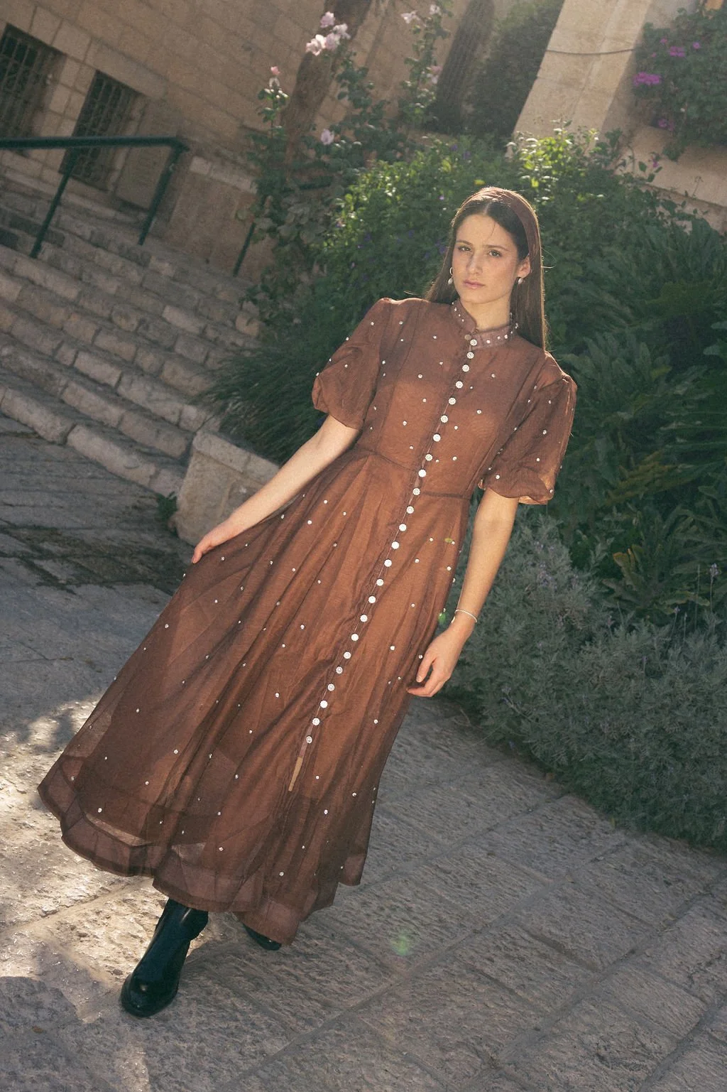 A woman in a brown dress with pearl embellishments standing outdoors on a stone-paved area with greenery and stone steps in the background.