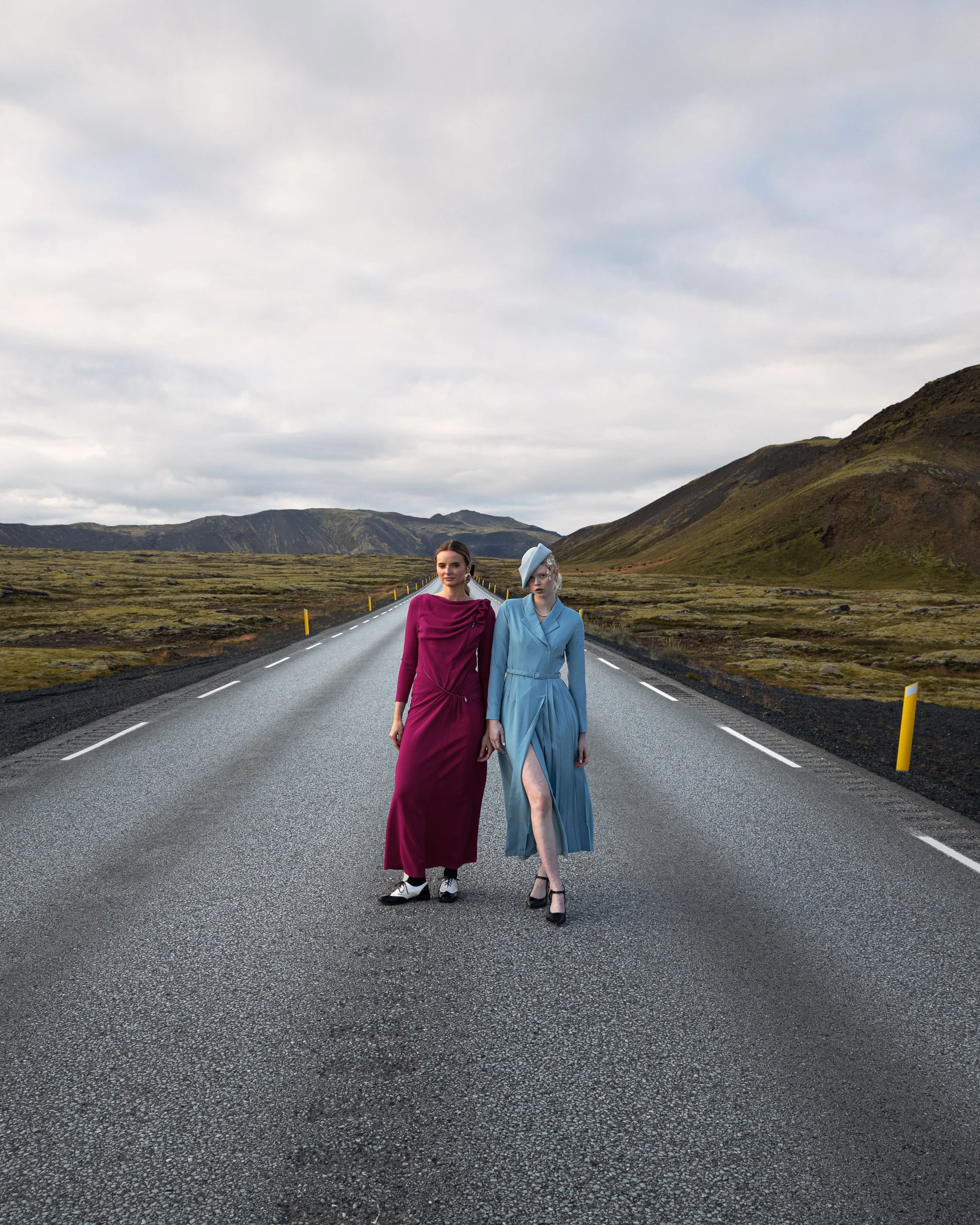 Two women in fashionable dresses walking hand in hand on an empty road through a mountainous landscape with cloudy skies.