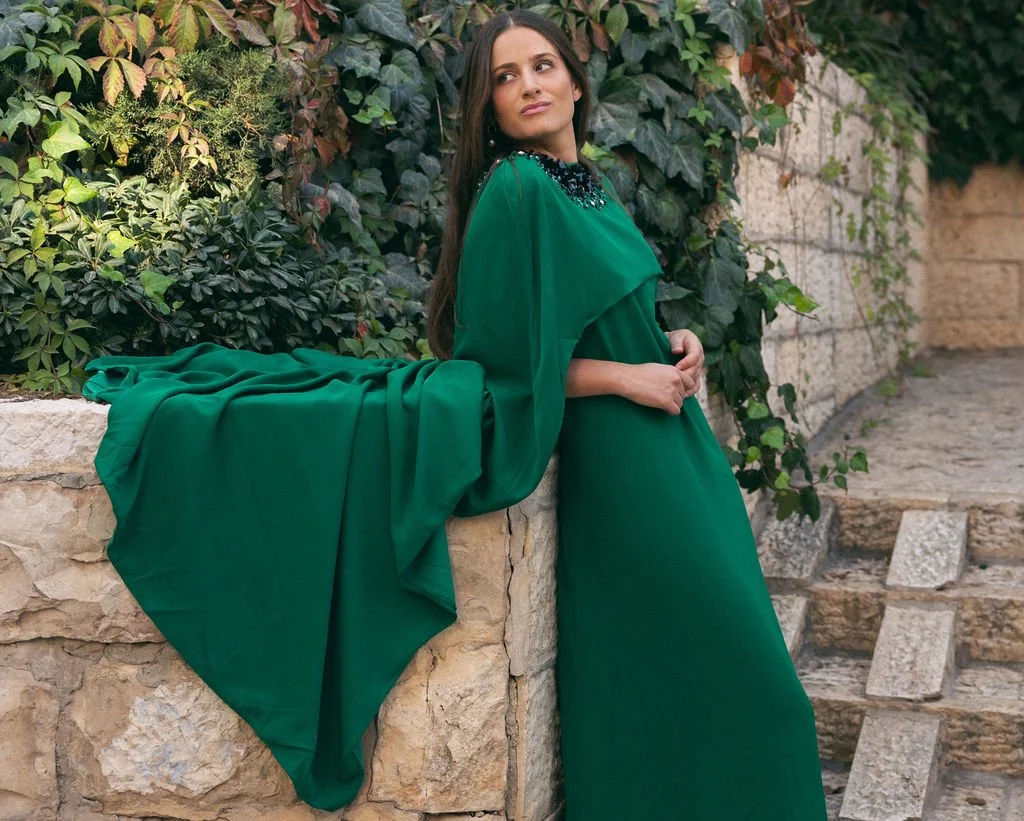 Woman in a long green dress with a black embellished collar, standing outdoors next to a stone wall with ivy and plants.