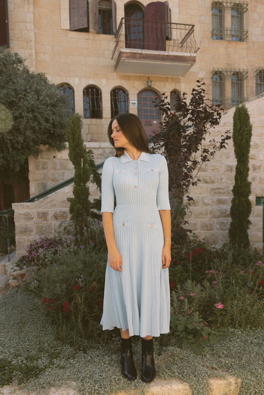 A woman in a light-colored, collared, button-up dress with pockets and black boots stands in a garden with flowering plants, in front of a stone building with multiple windows and balconies.