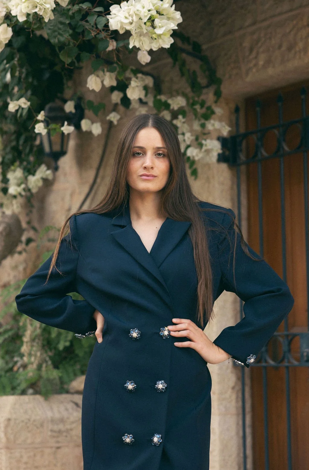 A young woman with long brown hair wearing a navy blue blazer dress adorned with decorative buttons, standing outdoors with green foliage and white flowers in the background.