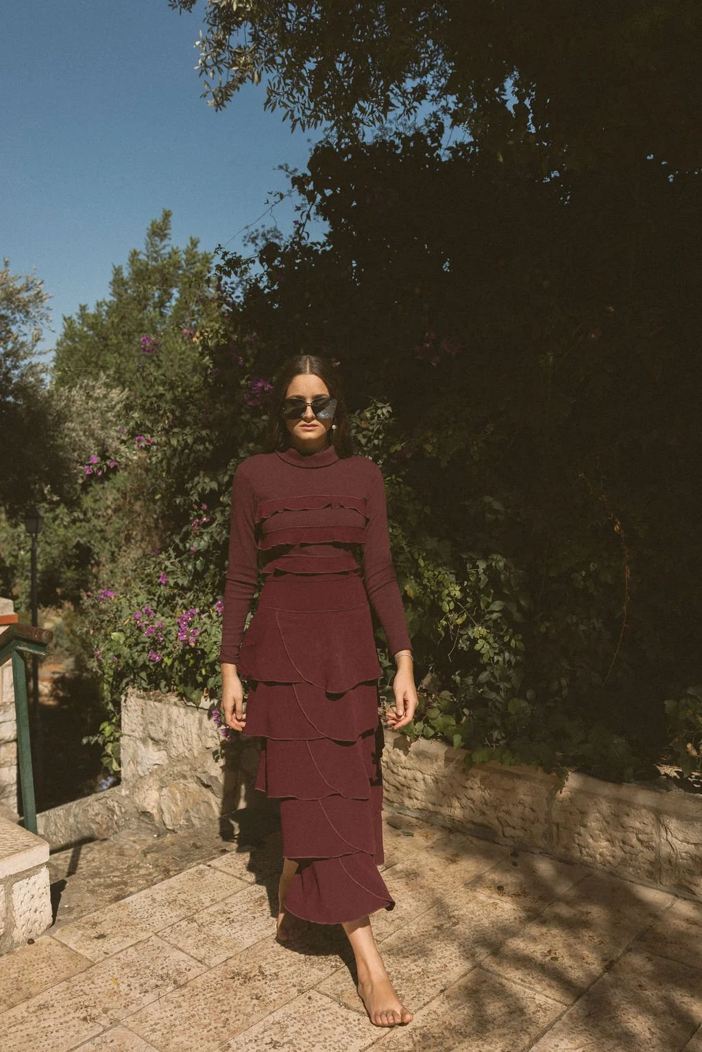 A woman wearing a maroon layered dress and sunglasses walks barefoot on stone pavement outdoors, with lush green trees and purple flowers in the background, under a clear blue sky.