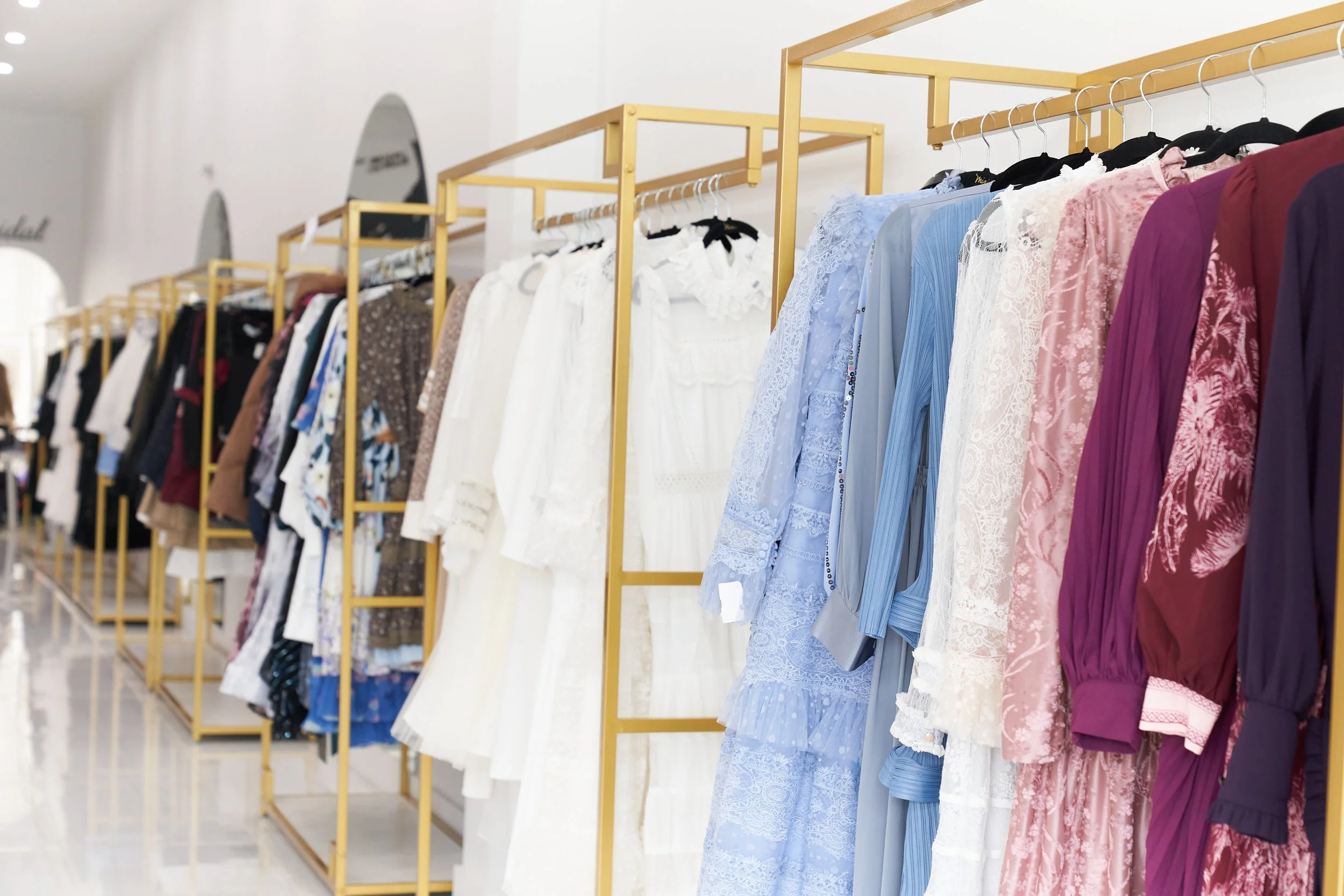 Clothing displayed on gold-colored racks in a store, with a variety of dresses and tops in different colors including white, blue, pink, and purple.