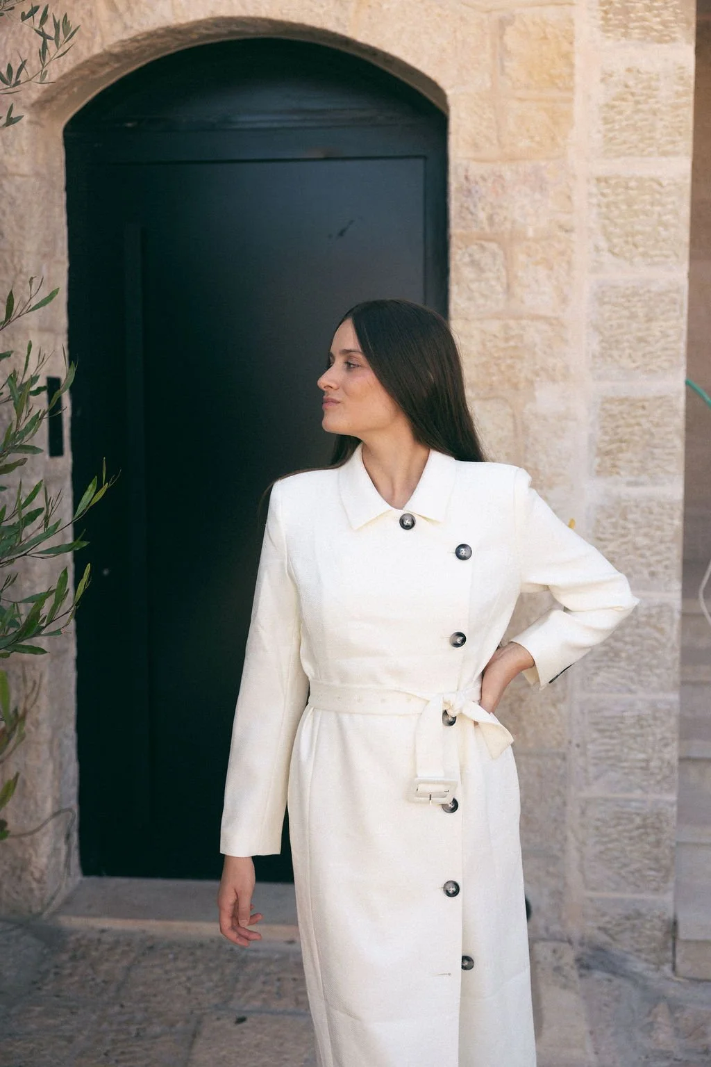 Woman with long dark hair in a white double-breasted coat with black buttons standing outdoors near a stone building and a black door.