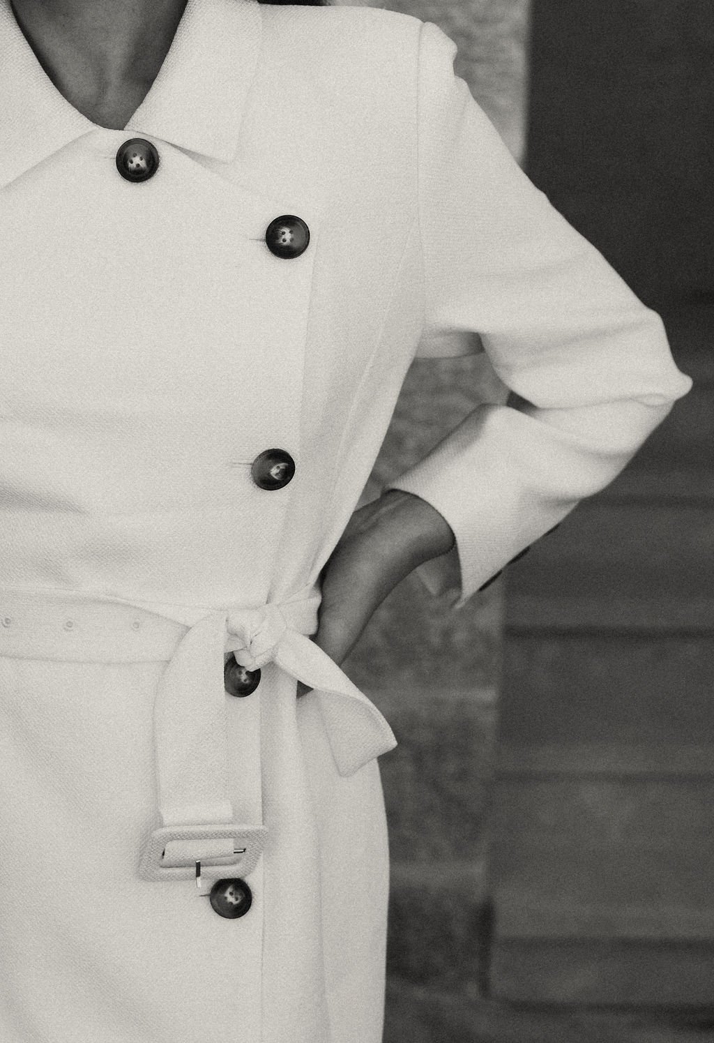 A person wearing a white double-breasted coat with black buttons and a waist belt, posing with one hand on the hip. The image is in black and white.