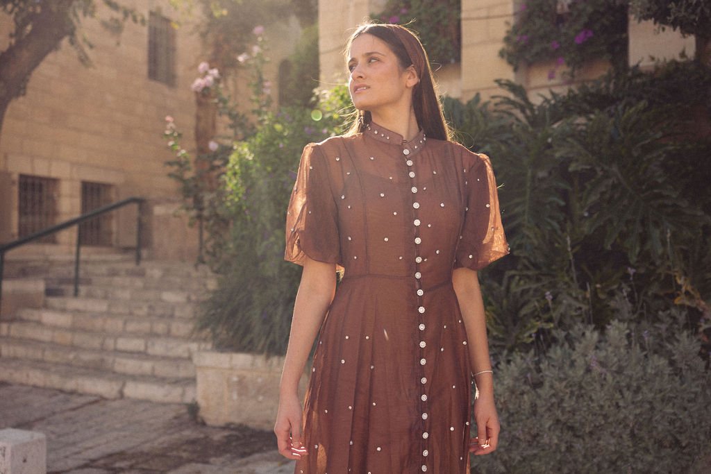 Young woman standing outdoors in sunlight, wearing a brown dress with pearl accents, surrounded by greenery and stone buildings.