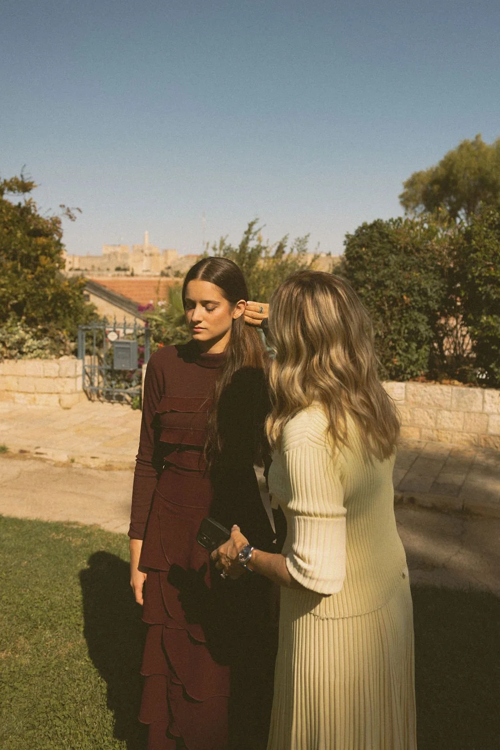 Two women standing outdoors on a sunny day, engaged in conversation, with a stone wall and trees in the background.