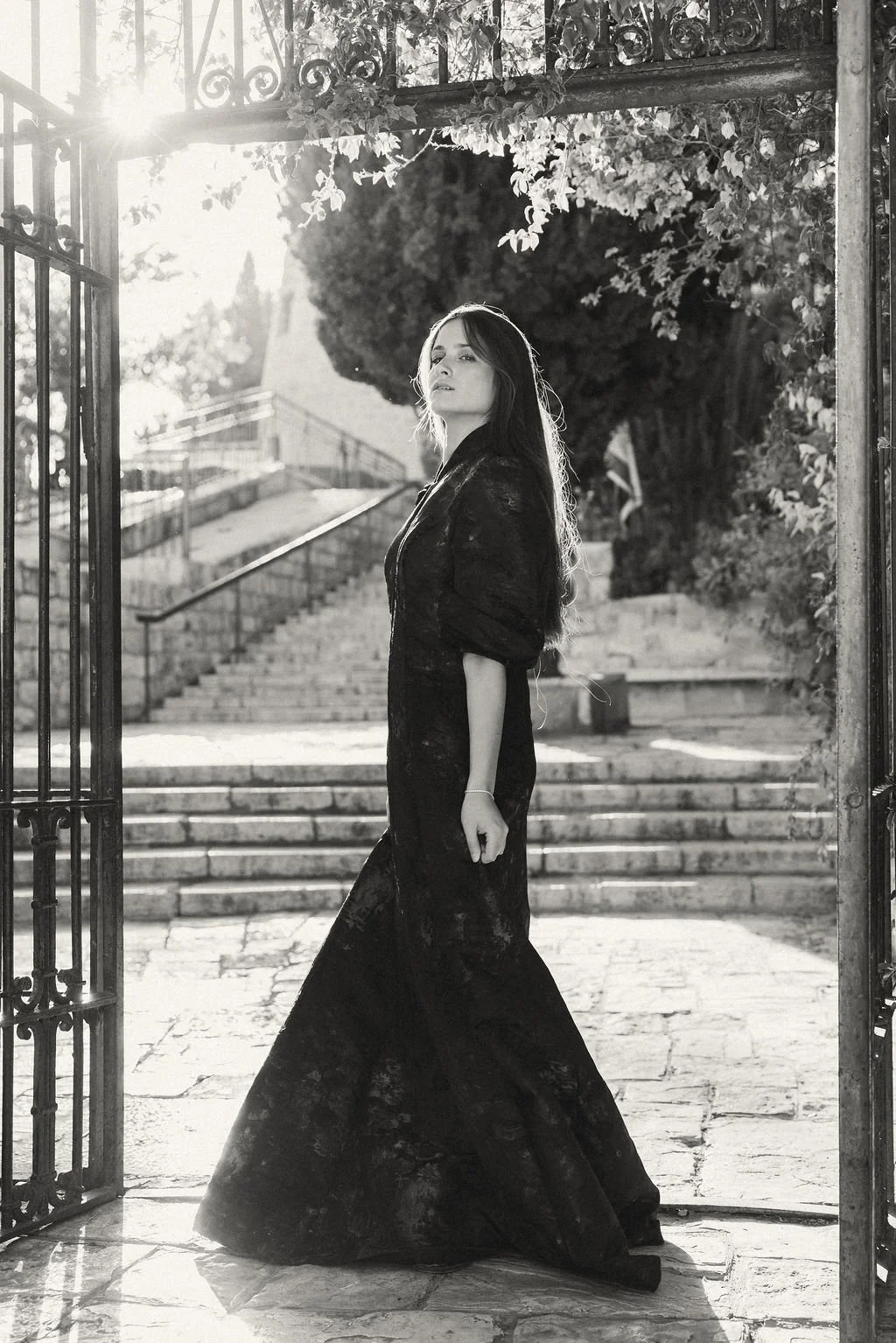 A woman in a dark dress stands through an open wrought iron gate, with stairs and trees in the background, sun shining from the left.