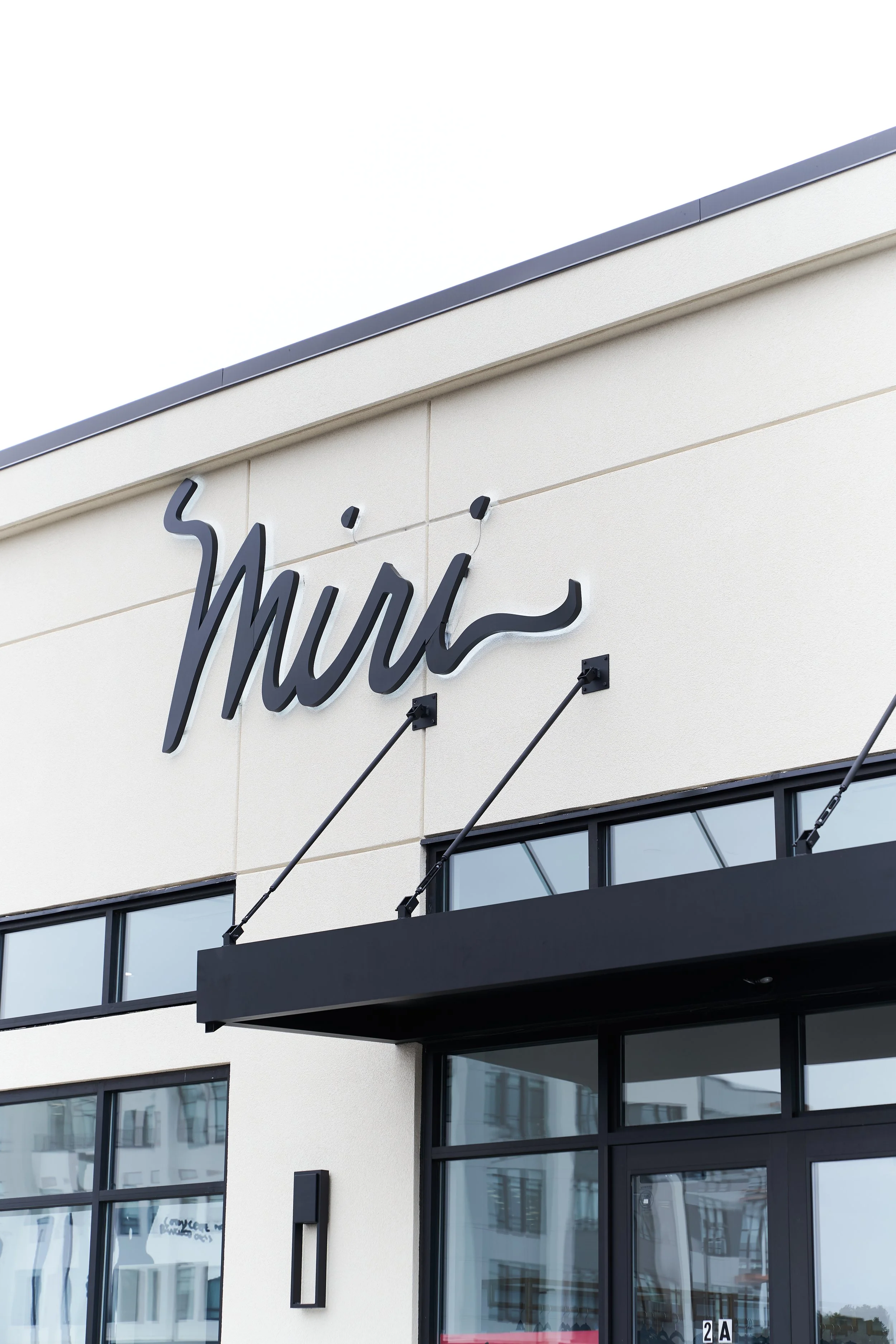 Exterior of a building with a sign that reads 'Mira' in black cursive letters, black awning, and glass windows.