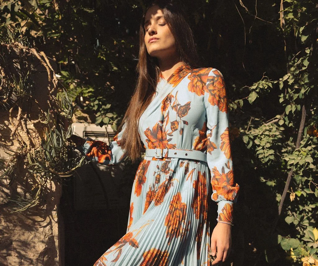 A woman in a floral dress with orange flowers and green leaves stands outdoors with her eyes closed, surrounded by greenery and shadowed foliage.