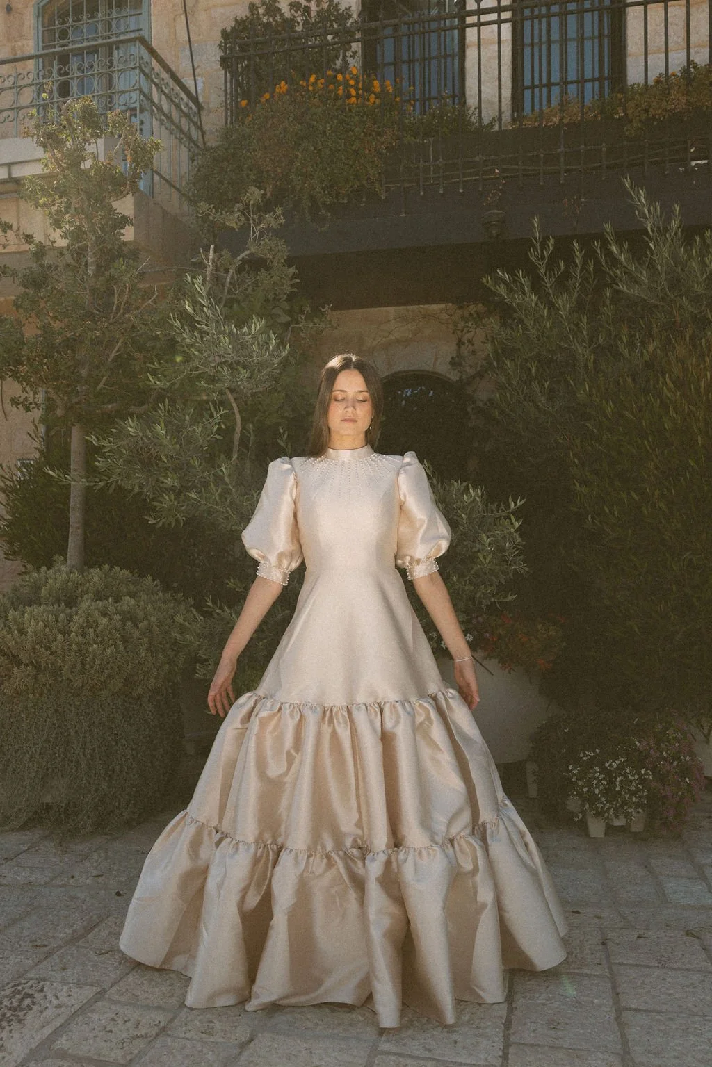 A woman wearing a long, elegant satin dress with puffed sleeves stands outdoors at night in front of greenery, with a building and balconies in the background.