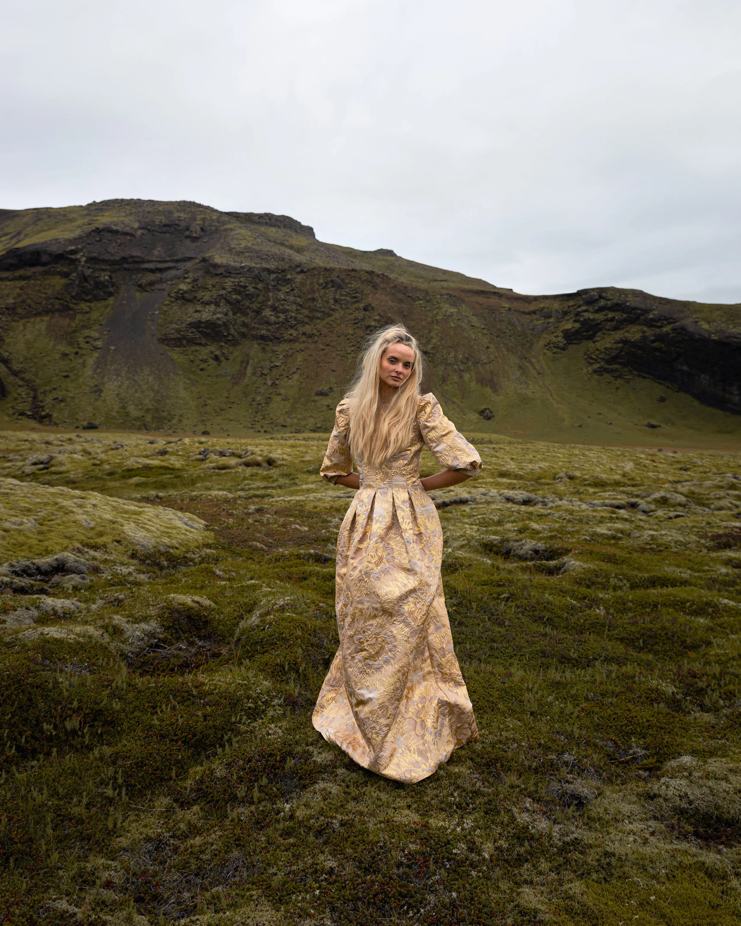 A woman in a gold and beige ornate gown standing in a green mossy landscape with mountains in the background and cloudy sky.