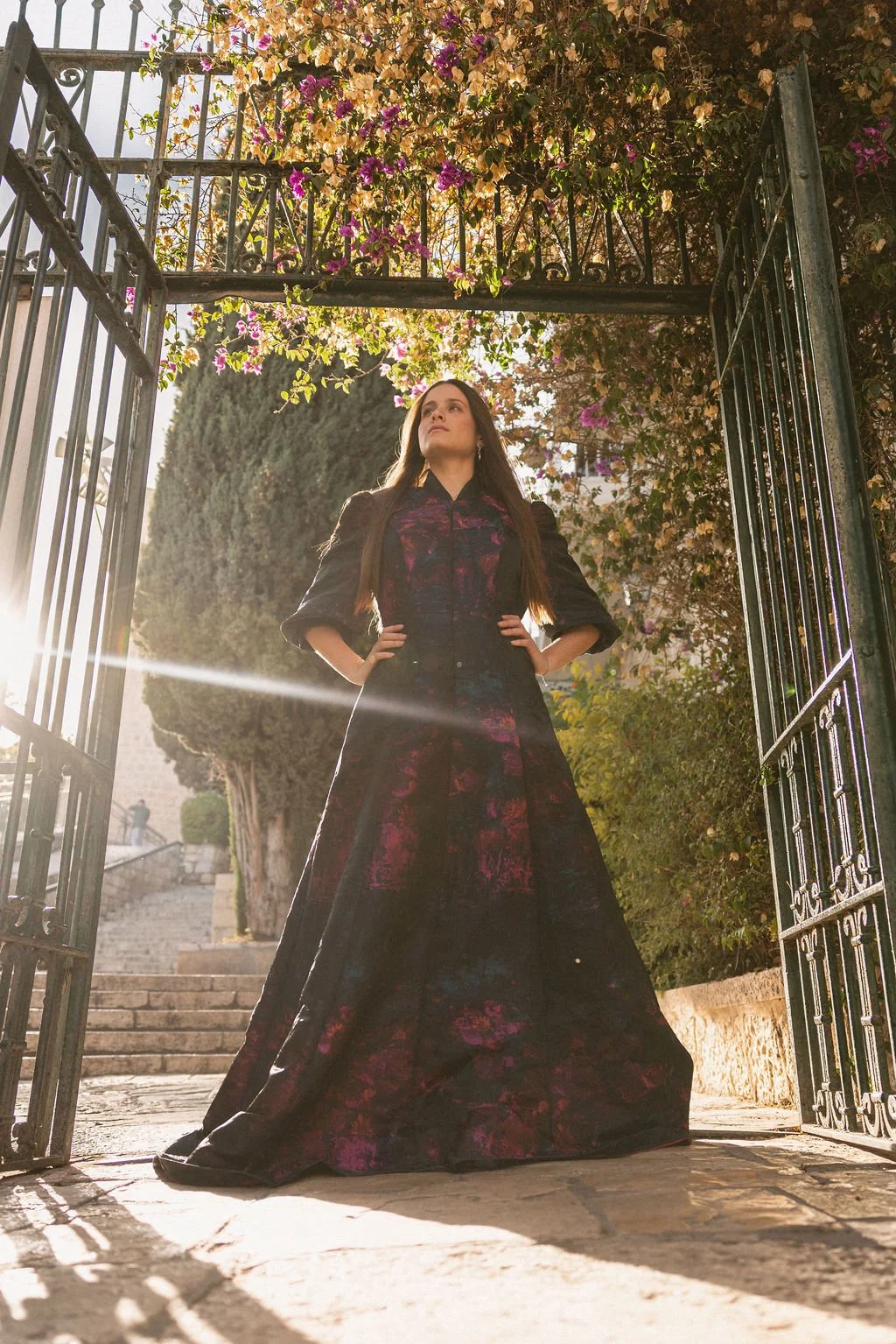 A woman in a long, dark floral dress standing with hands on hips at an open gate, surrounded by trees and flowers on a sunny day.