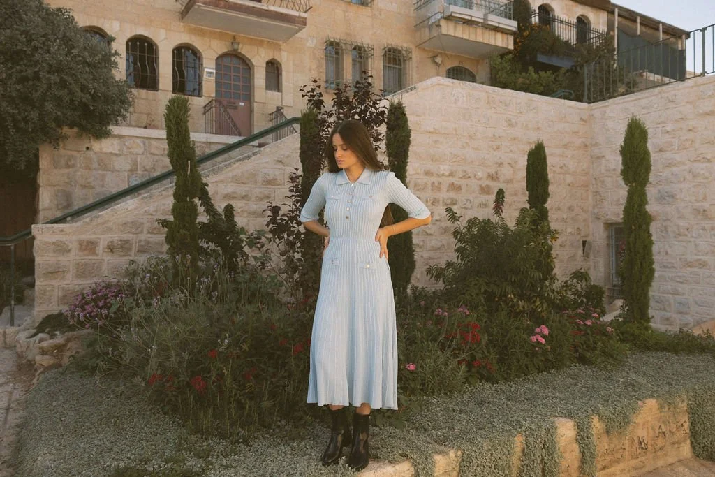 A woman with long dark hair wearing a light blue, vertically striped dress with buttons, standing in front of a stone wall and garden with flowers and tall trees.