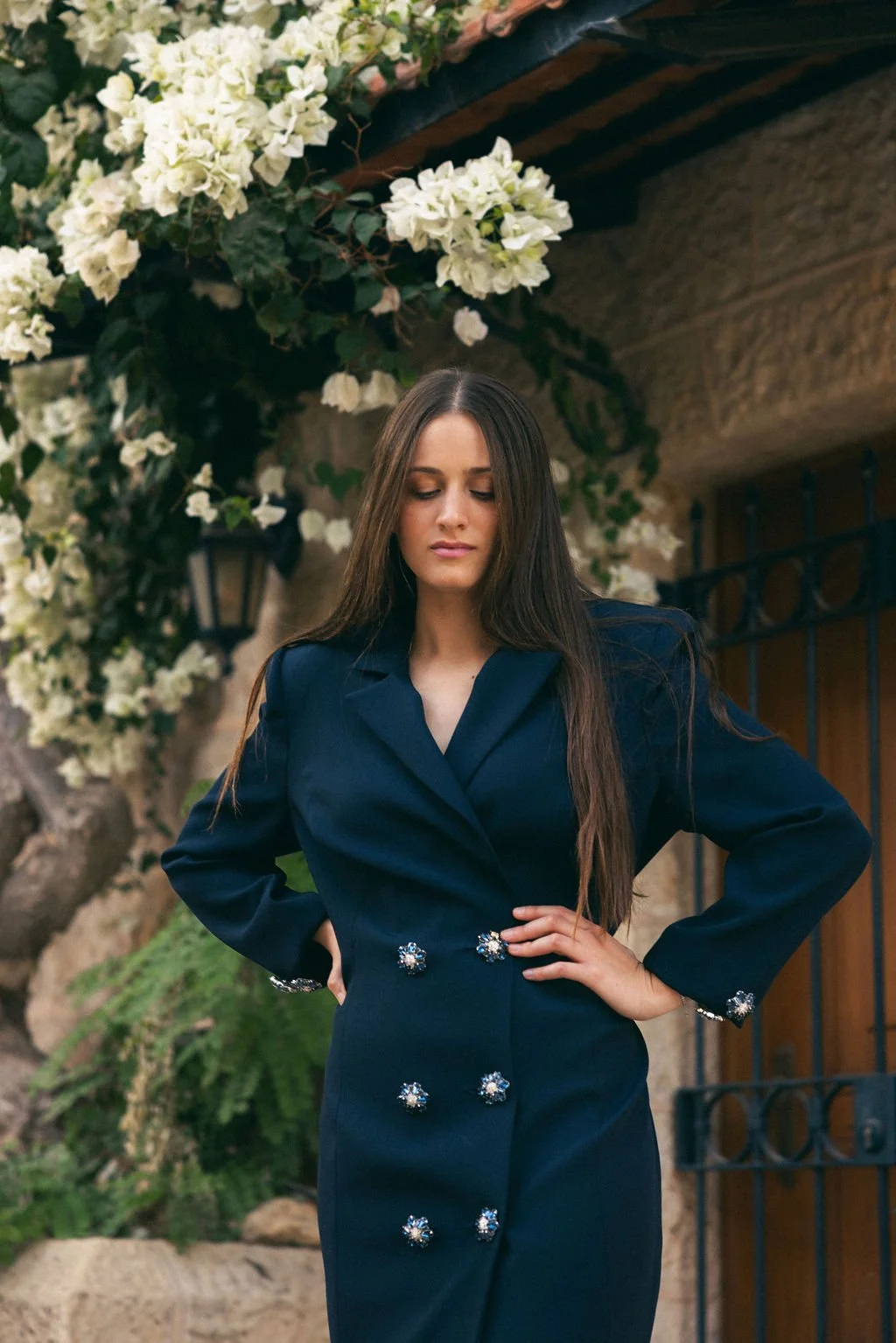 A woman with long brown hair wearing a dark blue dress with decorative buttons, standing outdoors in front of a building with a black iron gate and surrounded by white flowering plants.