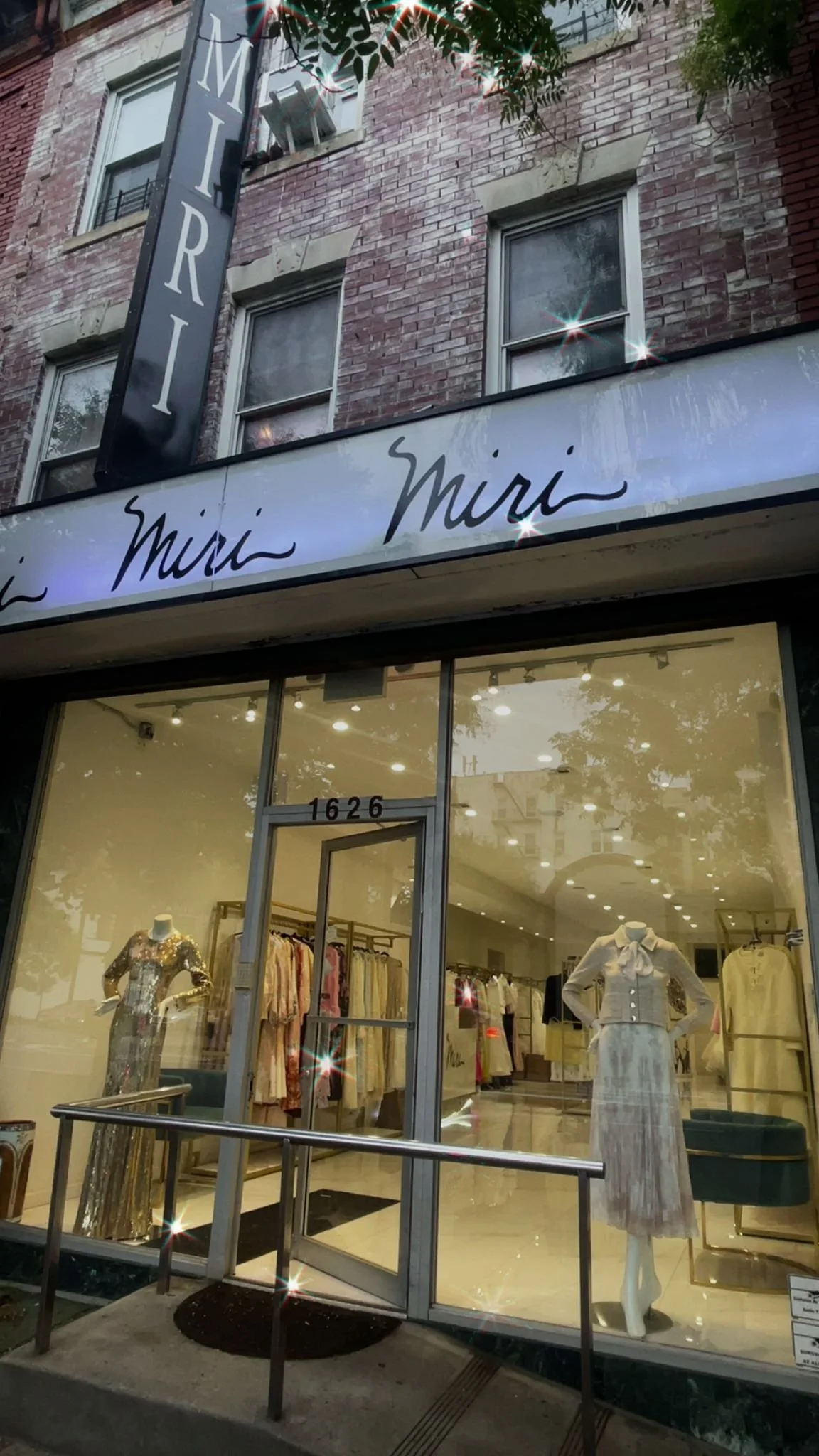 The storefront of Mimi, a clothing boutique, with large windows displaying mannequins dressed in stylish clothing inside. The store is housed in a brick building, and the sign above the entrance reads 'Mimi'.