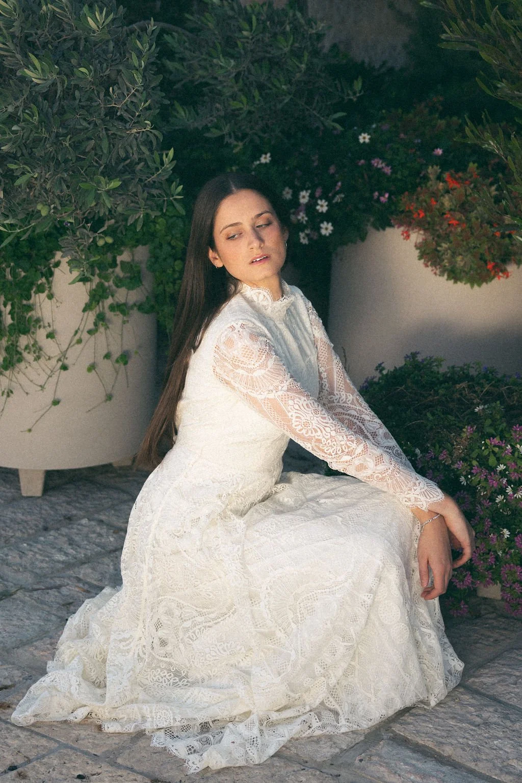 A woman with long dark hair wearing a white lace dress, sitting on a stone surface amidst potted plants and flowers.