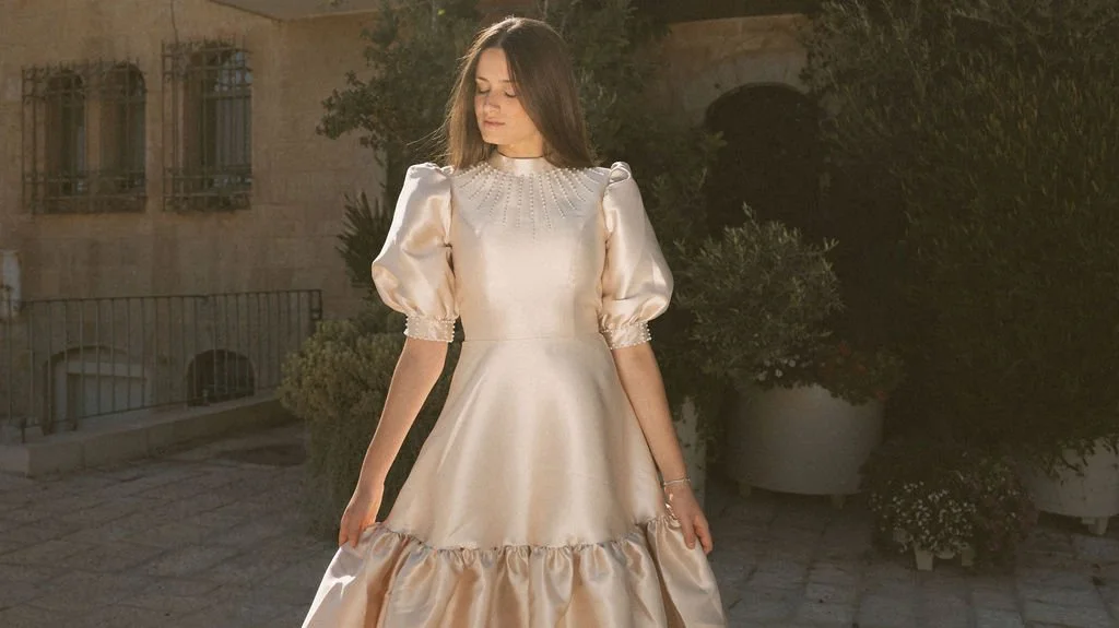 A woman wearing an elegant, shiny, cream-colored gown with puff sleeves and ruffle details stands outdoors with greenery in the background.