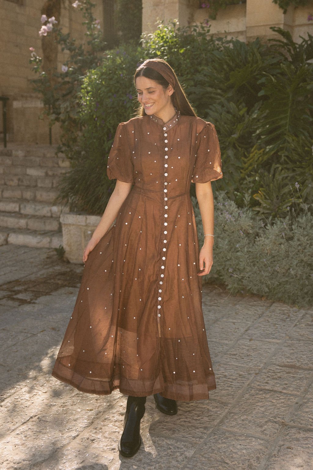 A woman with long brown hair wearing a brown sheer dress with white buttons and small white embellishments, standing outdoors on a stone pavement, smiling, with green foliage and stone steps in the background.