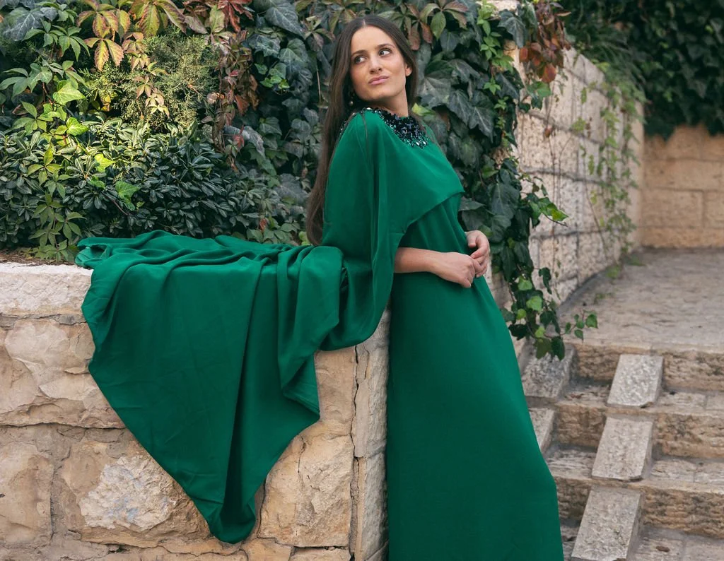 A woman in a flowing green dress with a beaded neckline standing against a stone wall with green foliage.