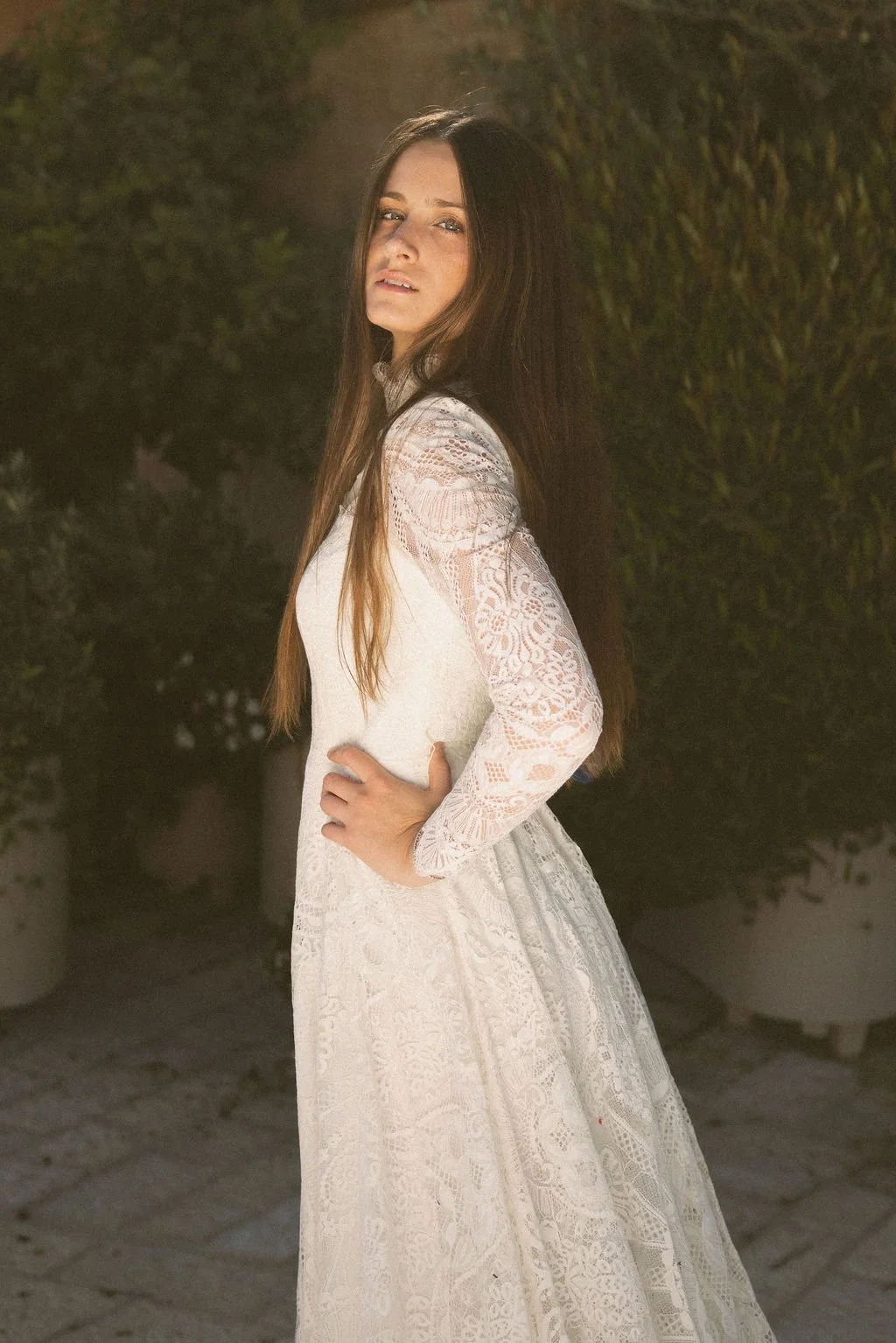 A woman with long brown hair wearing a white lace dress, standing outdoors near a hedge at night.