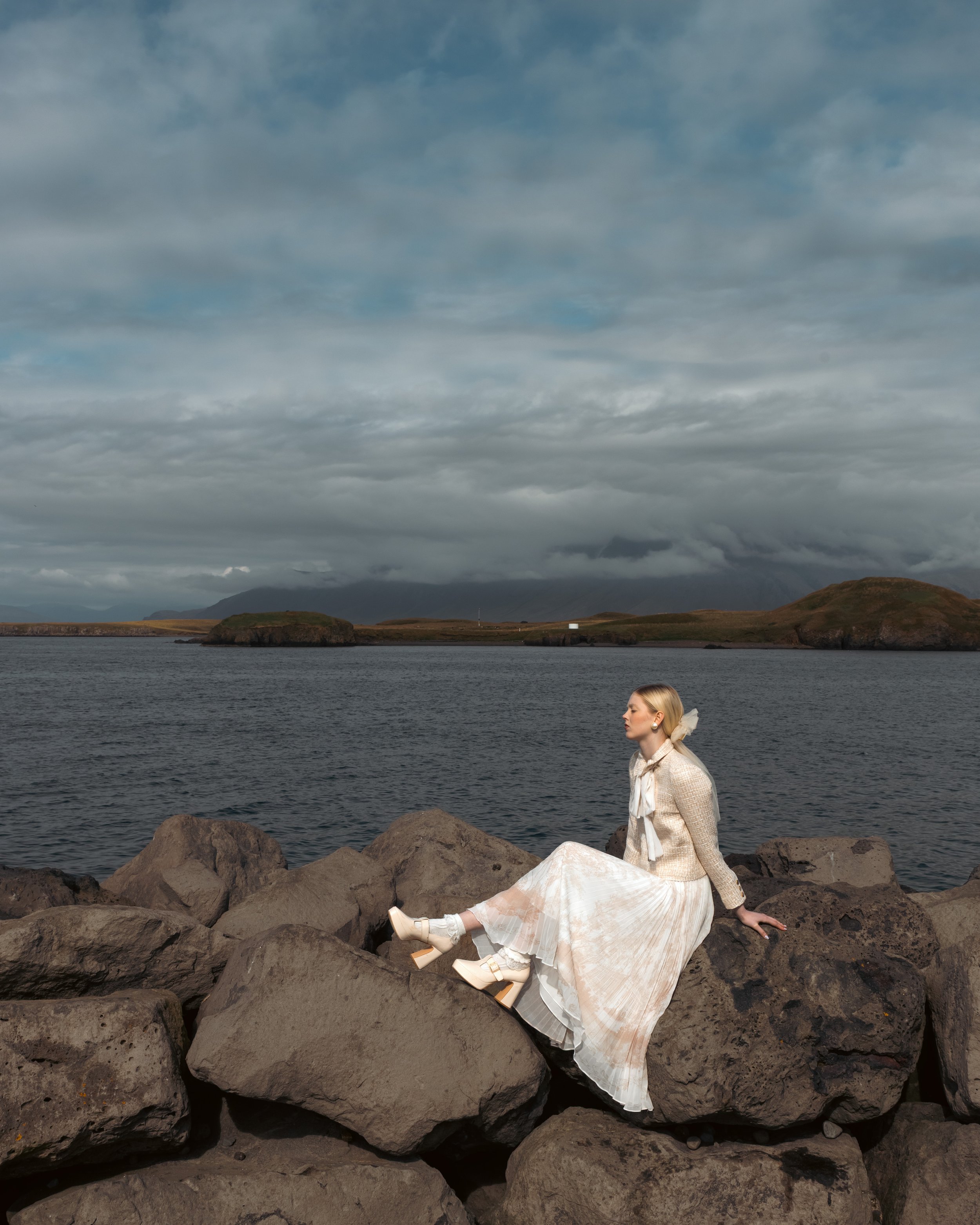 A woman in a light-colored dress and sweater sitting on rocks by a body of water, with land and overcast sky in the background.