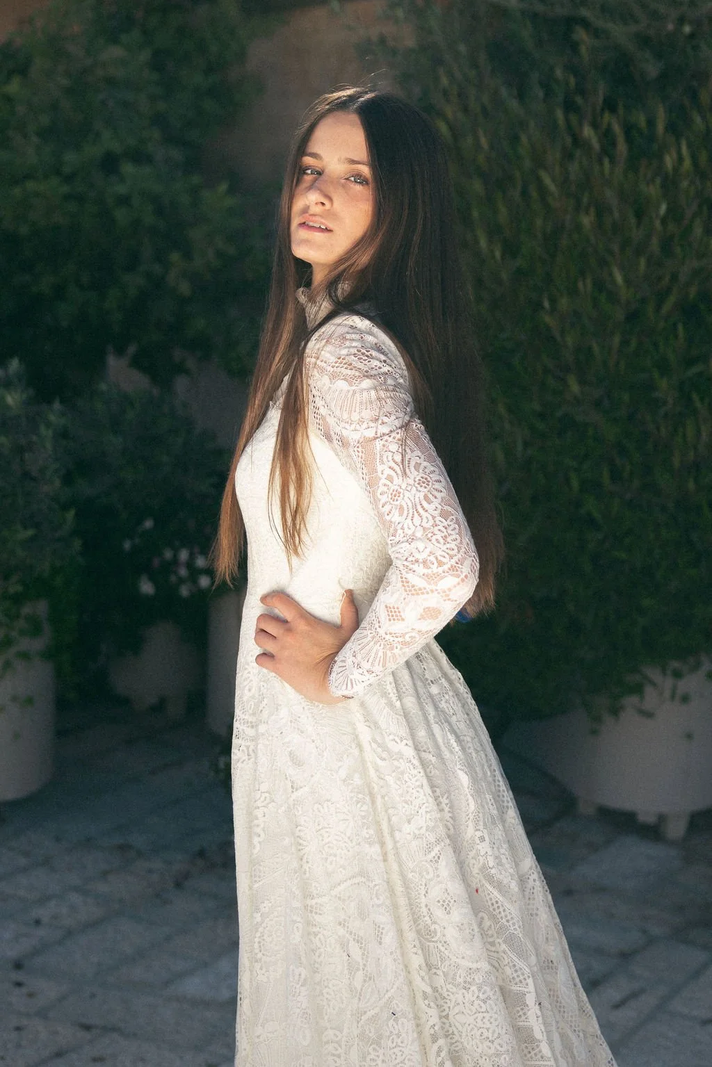 A woman with long brown hair wearing a white lace dress, standing outdoors with greenery in the background.