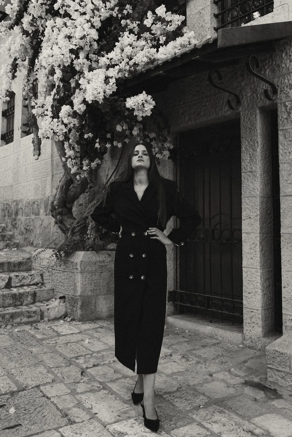 A woman in a black dress and heels standing on a cobblestone street at night with a flowering tree and stone building in the background.