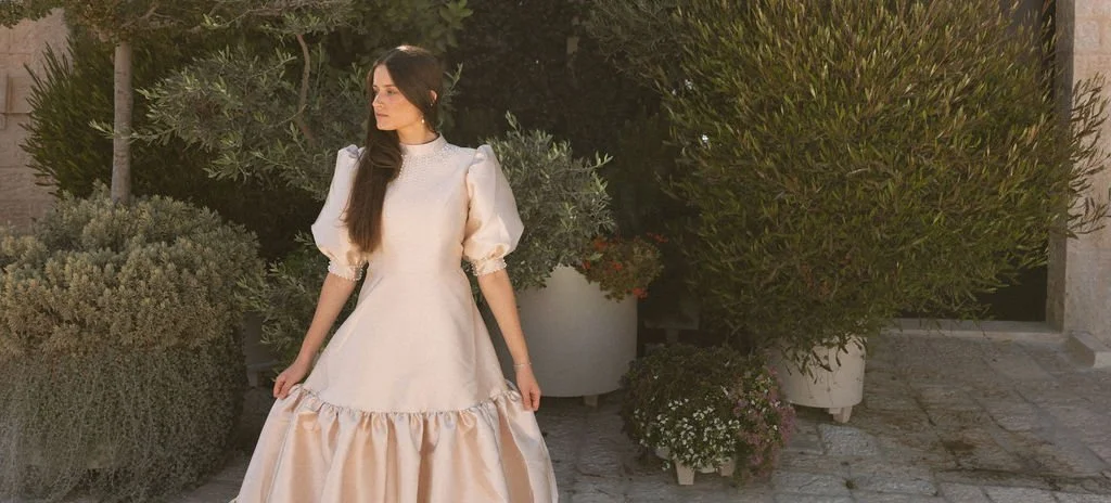 Woman in a vintage-style white dress standing among potted plants outdoors.
