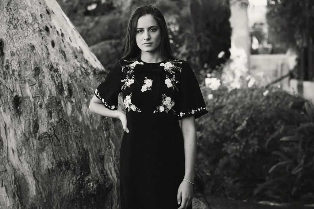 A woman in a dark dress with floral embroidery standing outdoors near a tree.