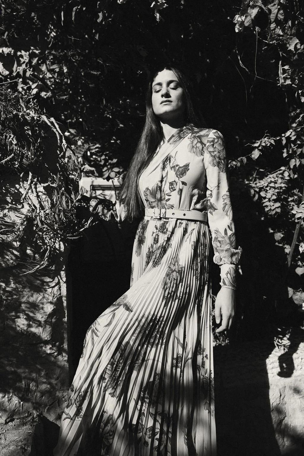 A woman with long hair wearing a floral patterned dress with a pleated skirt, standing outdoors among foliage, with sunlight and shadows creating dramatic lighting in black and white.