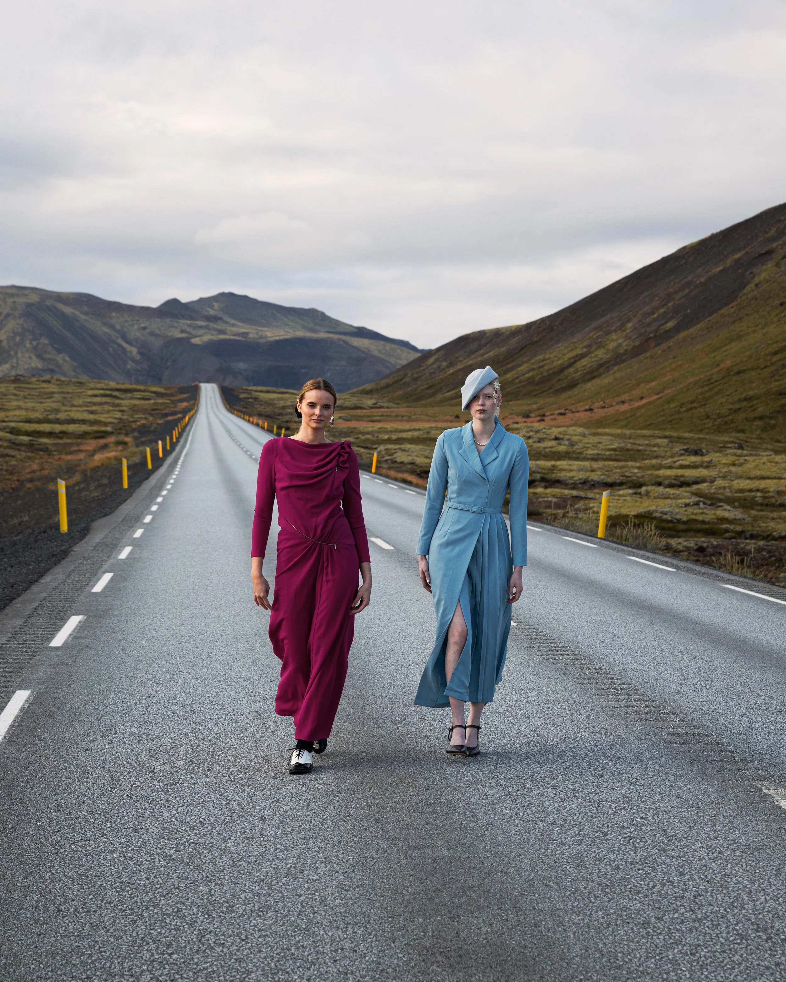 Two women in elegant dresses walking on a deserted road in a mountainous landscape with cloudy skies.