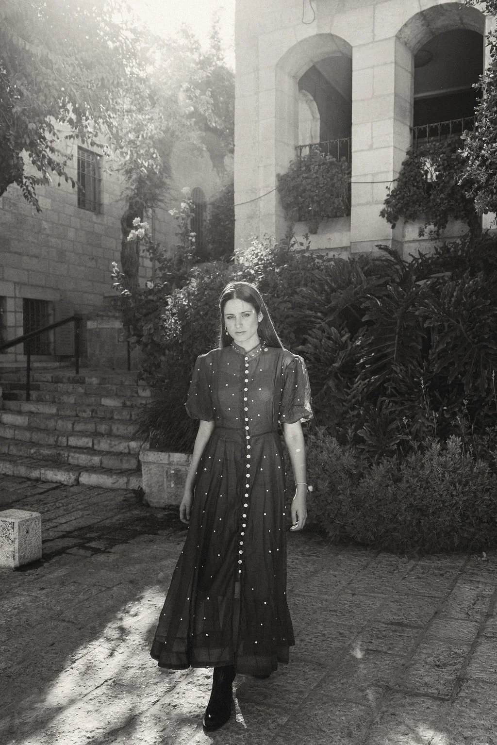 A woman in a long, dark, button-dress stands outdoors on stone pavement, with lush bushes and historical stone buildings with arched windows and balconies in the background, illuminated by sunlight.