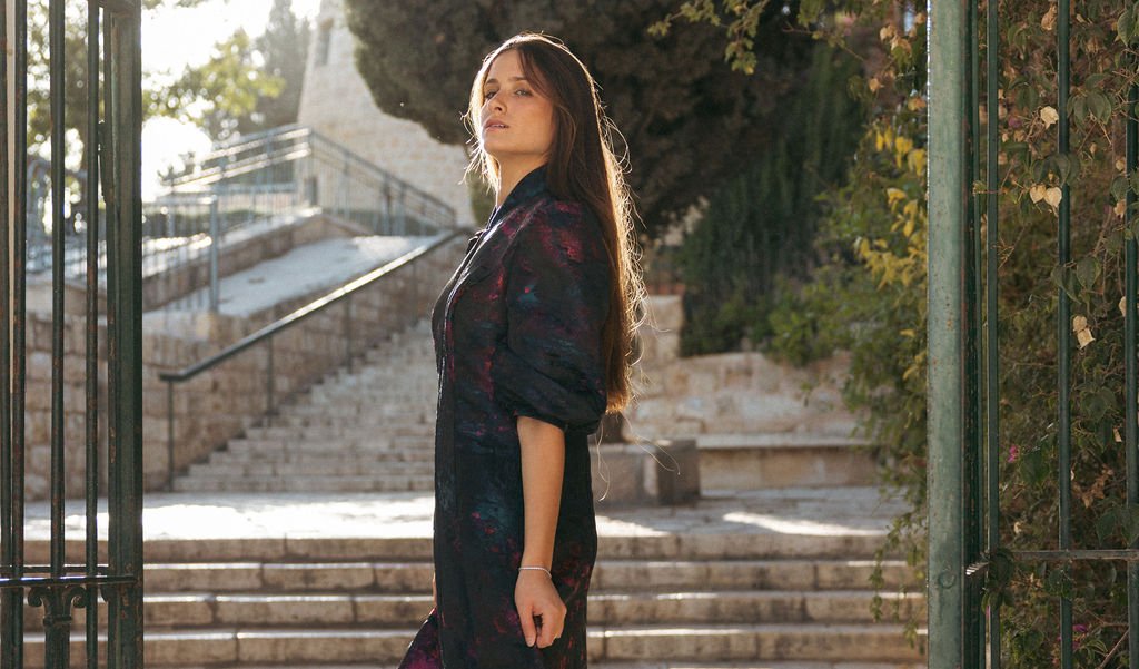 A woman with long dark hair standing near a wrought iron gate, with stone stairs and greenery in the background on a sunny day.