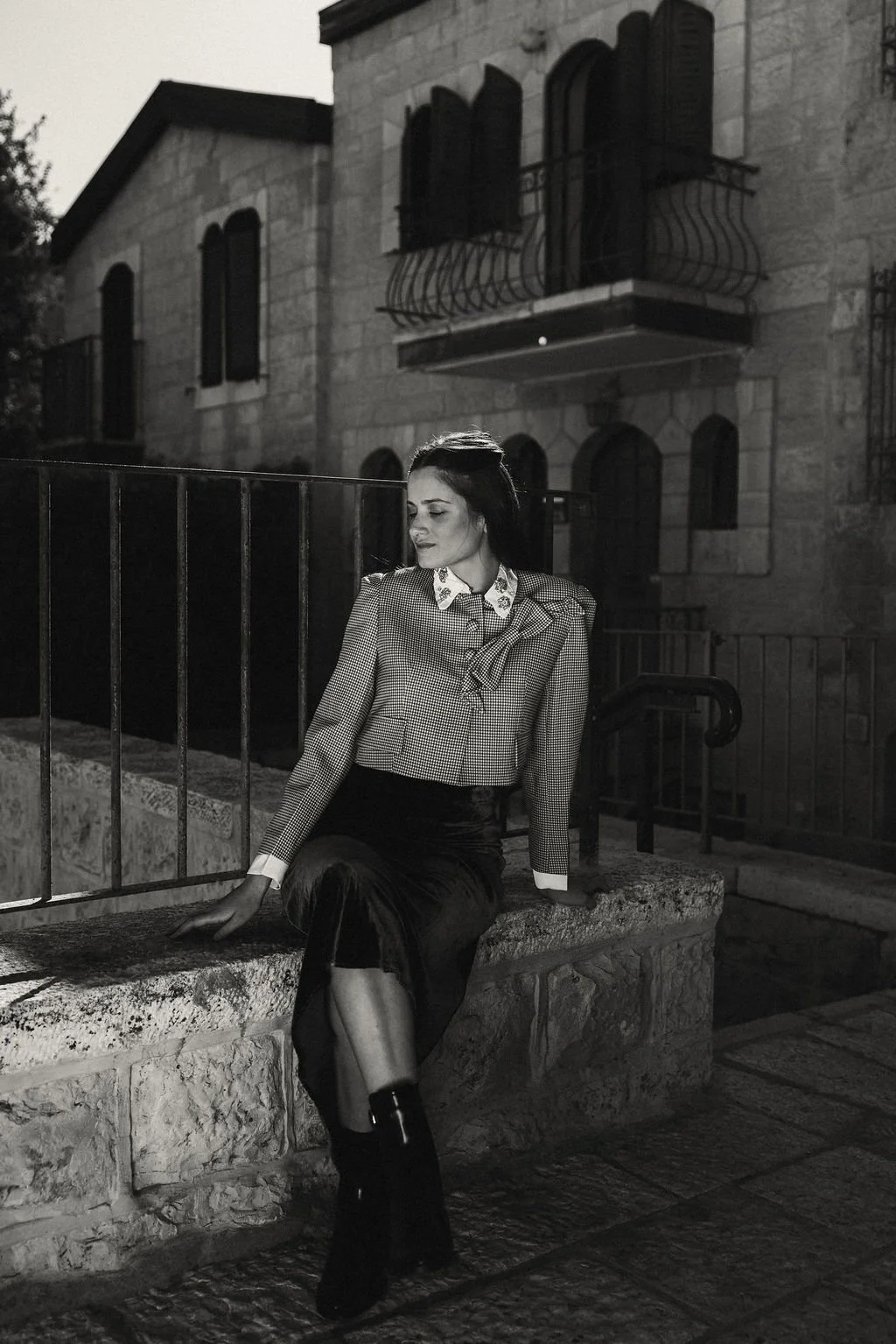 A woman dressed in a blazer and skirt, sitting on a stone ledge near a metal fence outside a building at dusk or night.