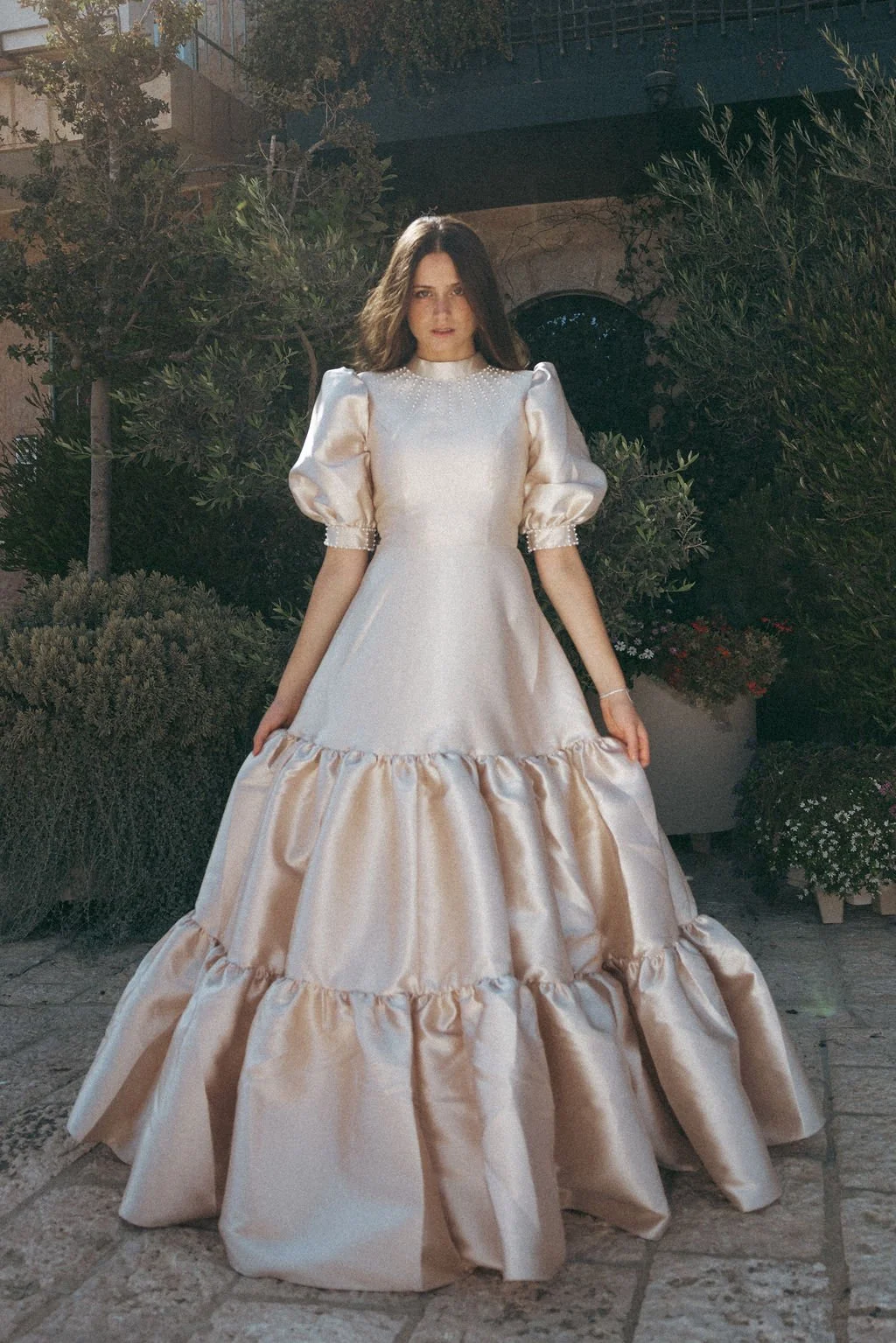 A woman in a long, elegant, satin gown with puffed sleeves and layered ruffles stands outdoors at night. She has medium-length dark hair and is surrounded by bushes and potted plants.
