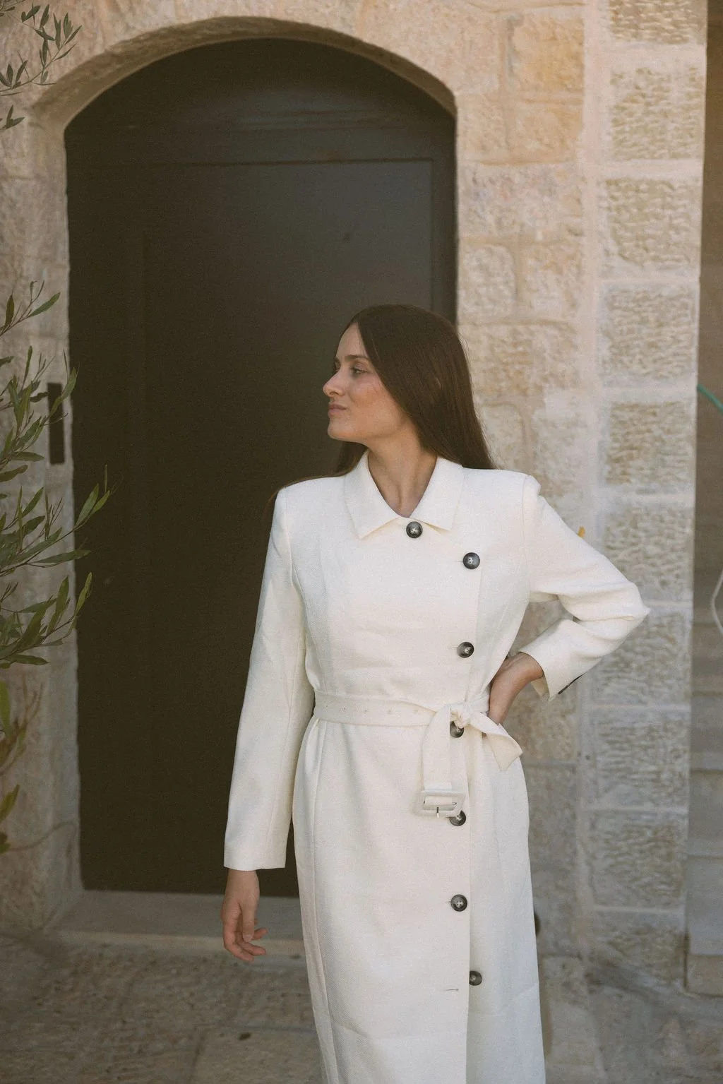 A woman in a white coat standing outside in front of a stone wall and black door.
