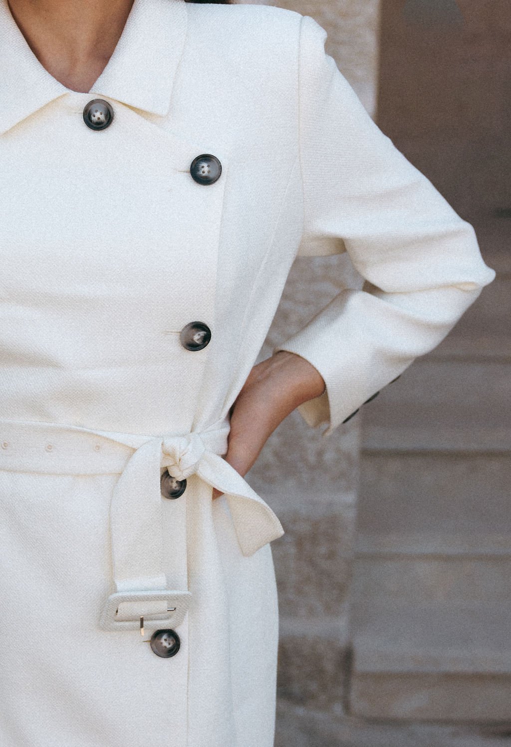 Close-up of a person wearing a white double-breasted coat with black buttons and a waist tie, standing with one hand in their pocket.