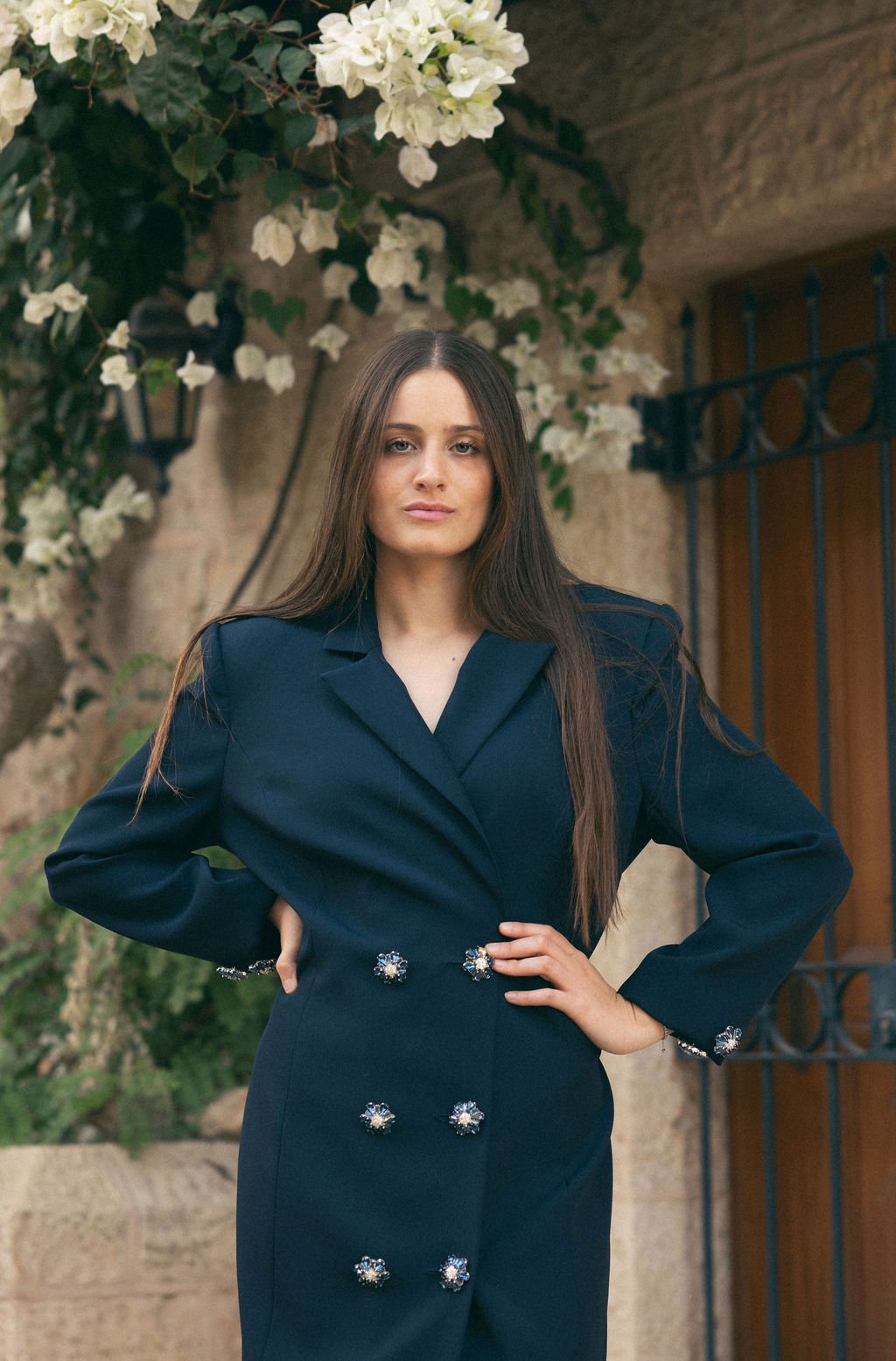 A woman with long brown hair wearing a dark blue blazer dress with decorative buttons, standing outdoors with her hands on her hips in front of a stone wall, greenery, and white flowers.