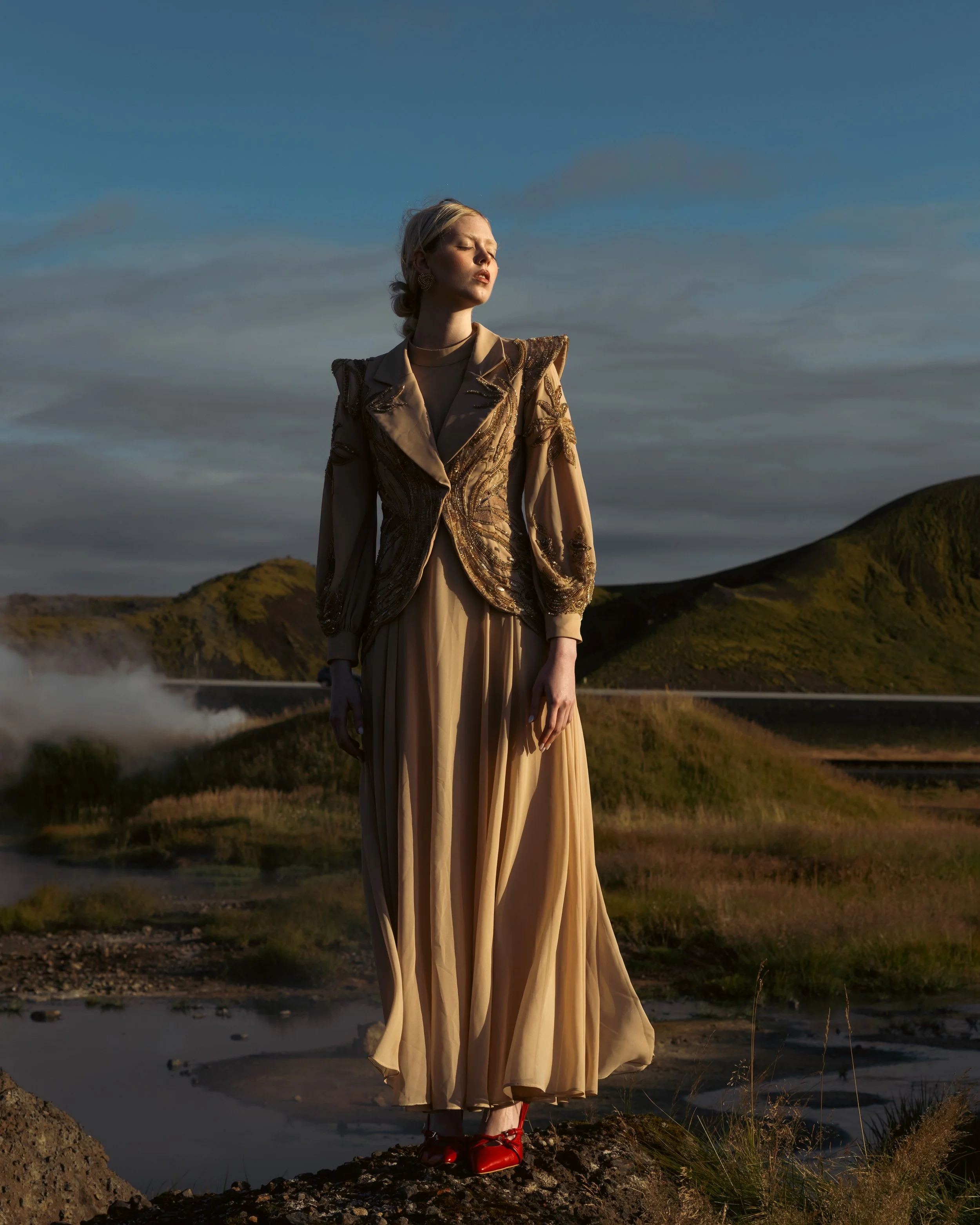 A woman in a gold and black embroidered jacket and a long beige dress with red shoes standing outdoors on rocky terrain with mountains and a partly cloudy sky in the background, illuminated by sunlight.